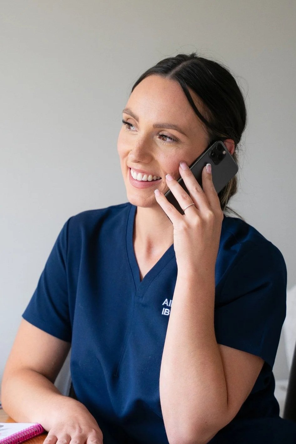 A young woman with dark hair, wearing navy scrubs, smiling while talking on a cell phone.