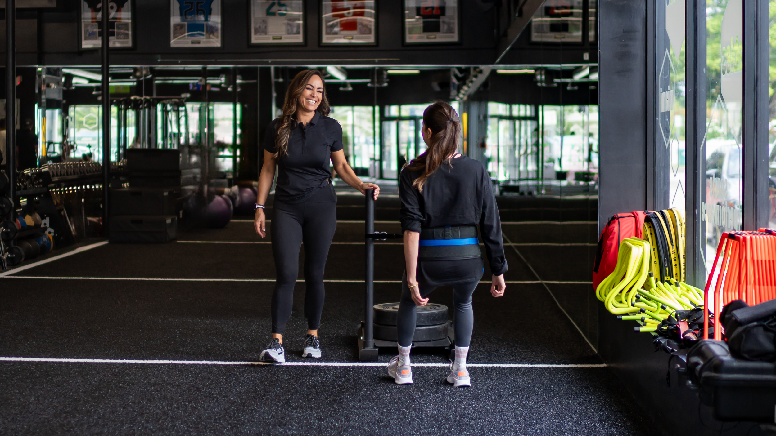 A trainer and a woman with a blue belt in a gym, standing near a sled for strength training, with workout equipment and bright yellow and orange training gear on the side.
