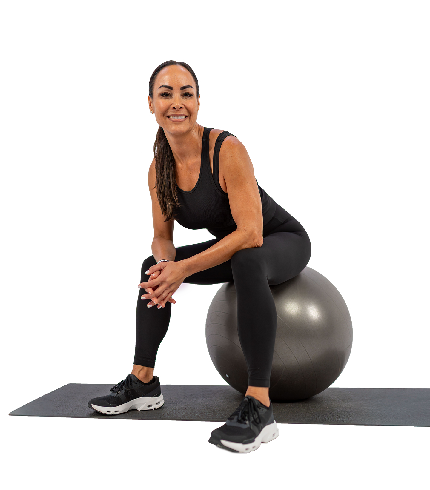A woman sitting on an exercise ball in workout attire, smiling, on a black exercise mat.