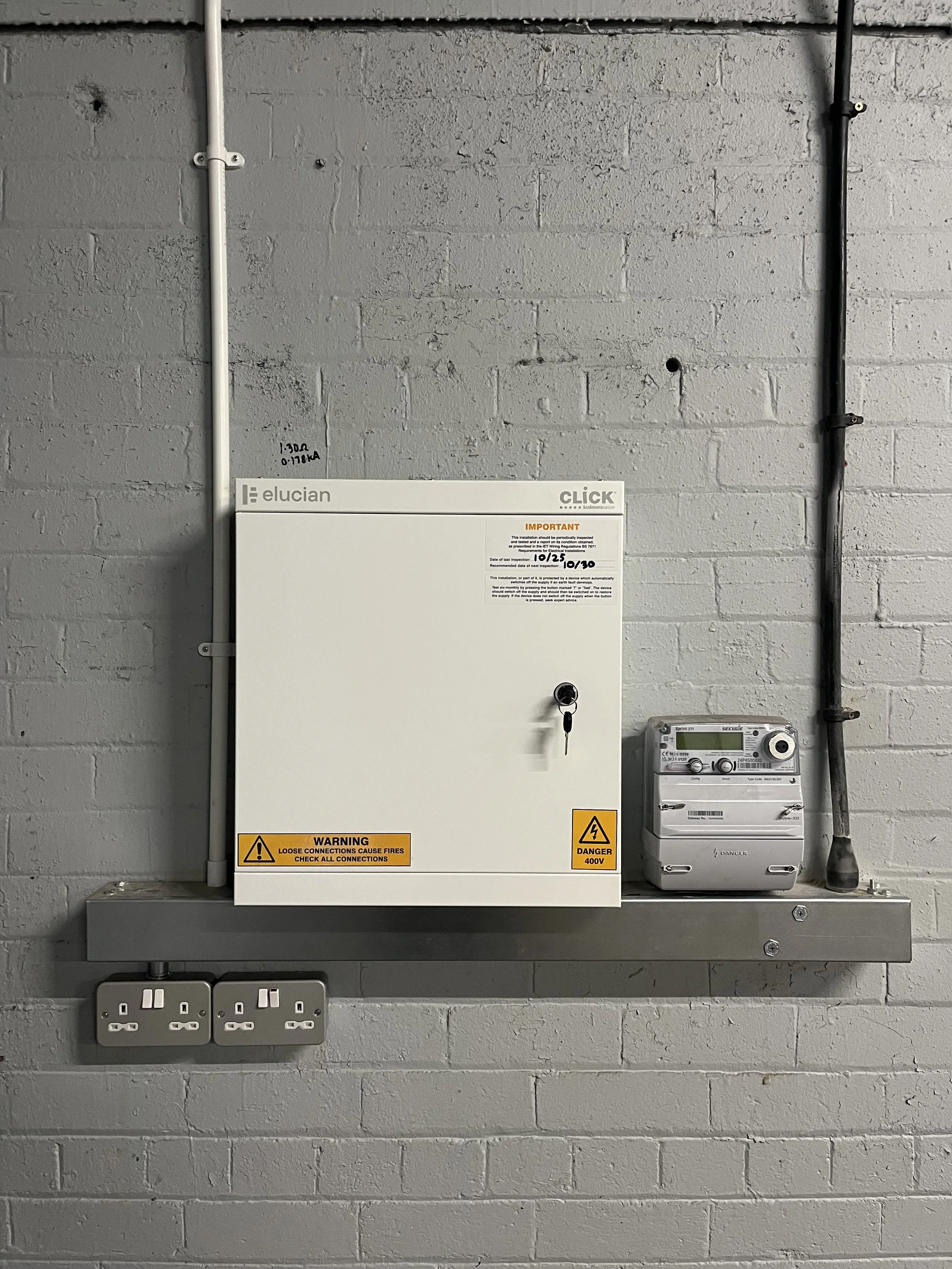 Electrical control panel mounted on a gray brick wall, with warning labels, two power outlets below, and electrical conduit pipes on the wall.