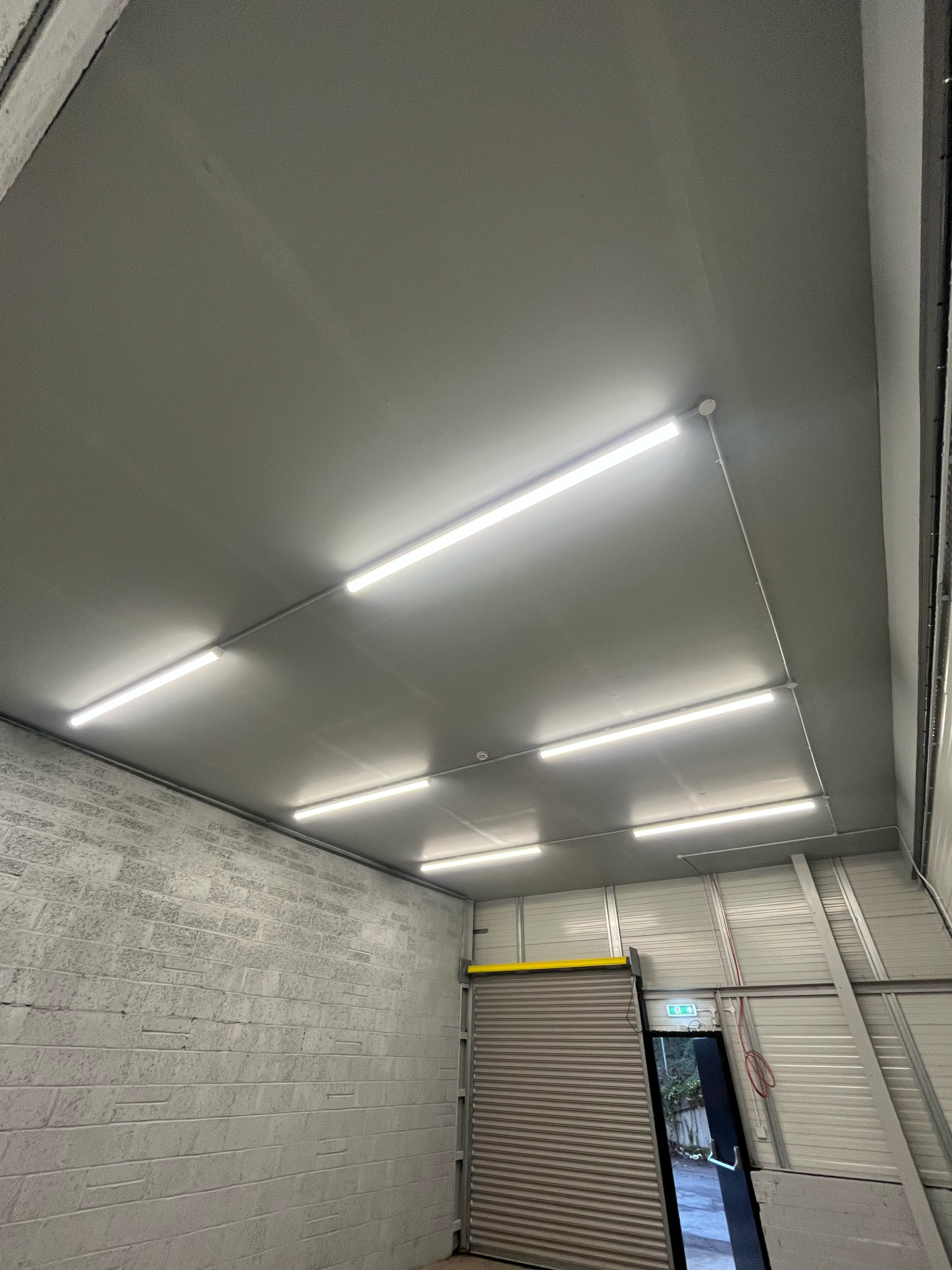 Interior view of a garage with white brick walls, ceiling with long fluorescent lights, roll-up metal door, and a small exit door with an emergency light above it.
