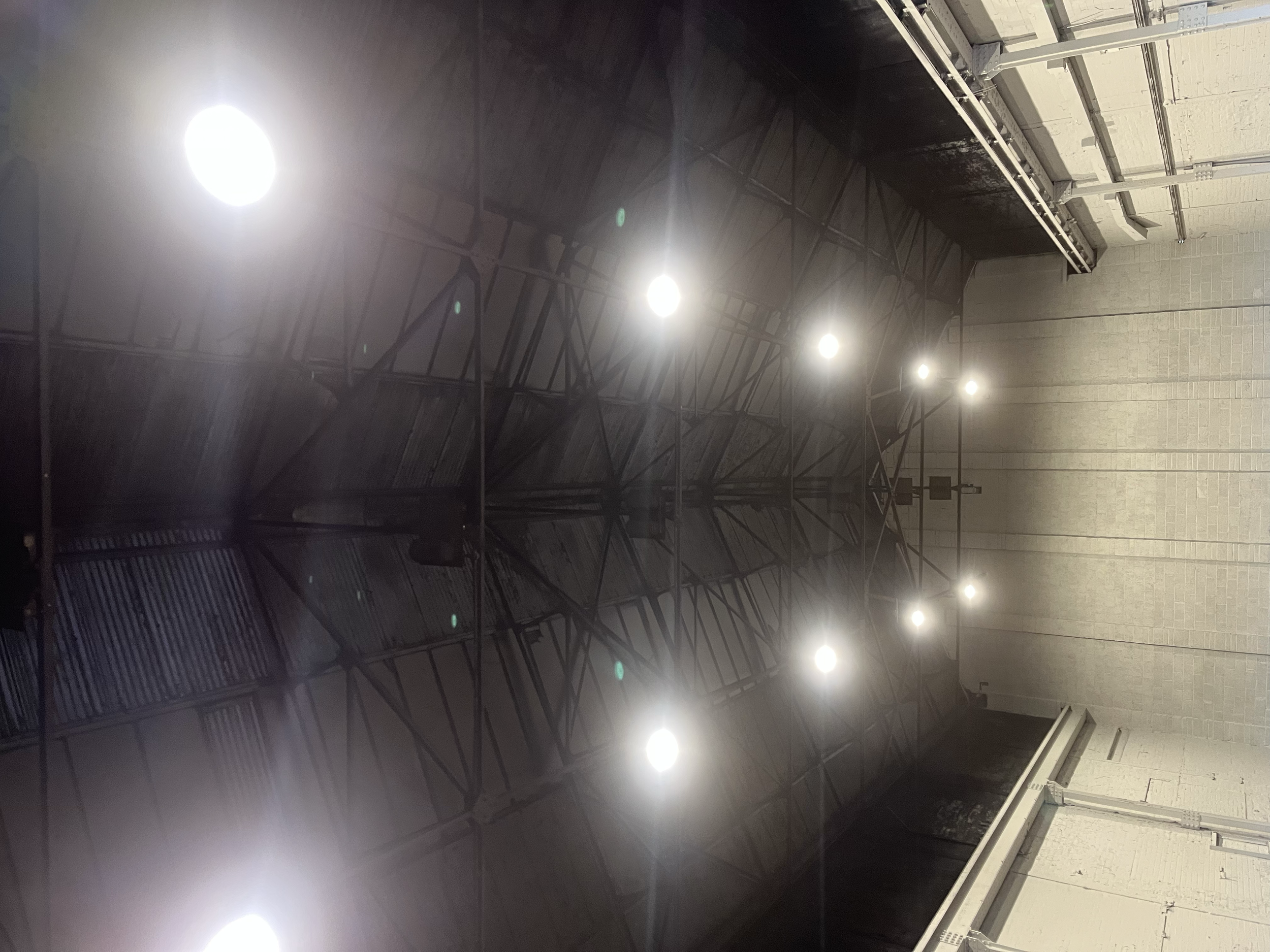 Indoor space with a high ceiling, bright ceiling lights, a dark wall, train tracks, and a platform or hallway with tiled walls.