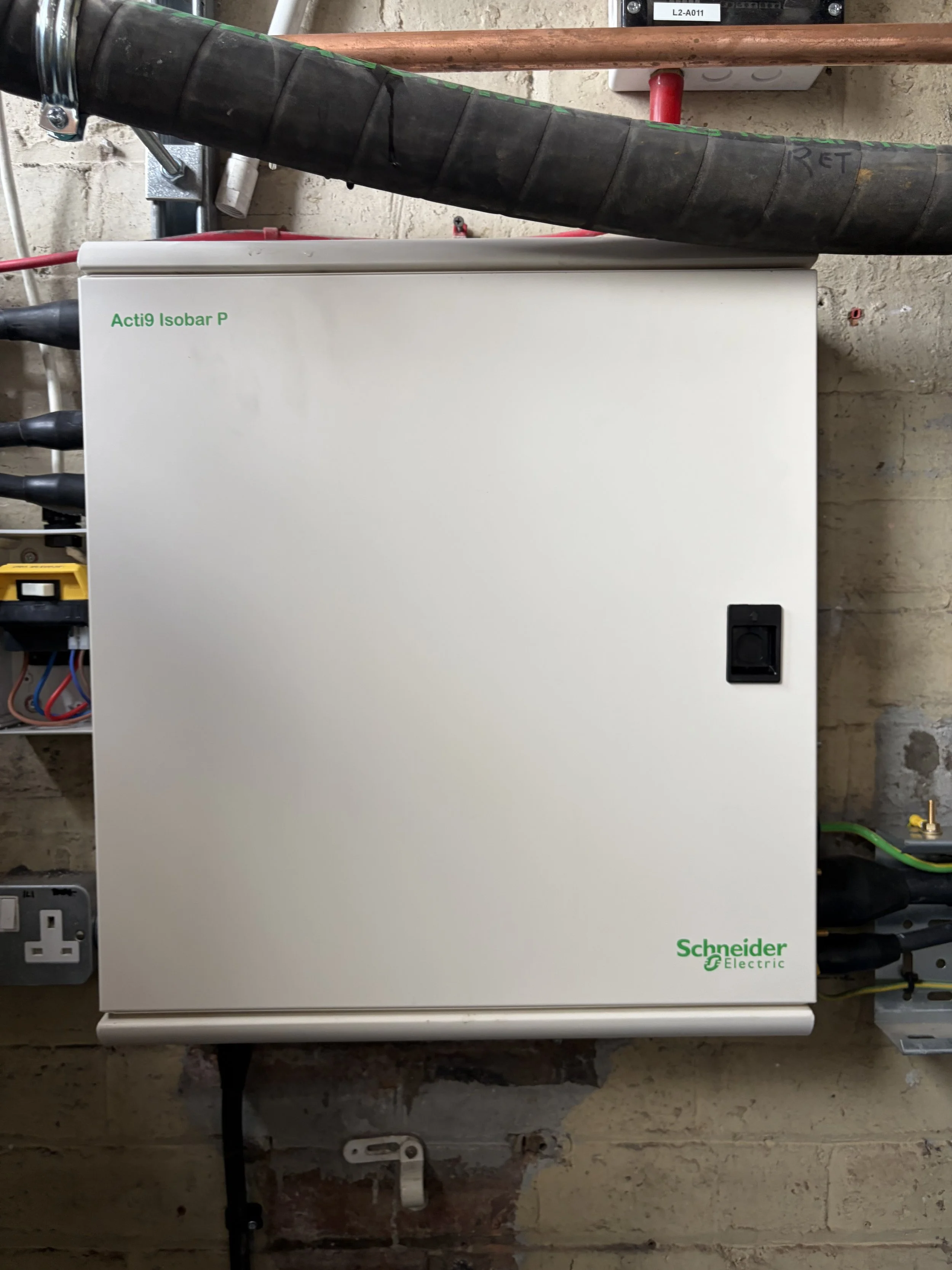 A white electrical control panel with green text that reads 'Acti9 Isobar P' and 'Schneider Electric' logo, mounted on a wall in an industrial or utility room, with various electrical wires and equipment nearby.