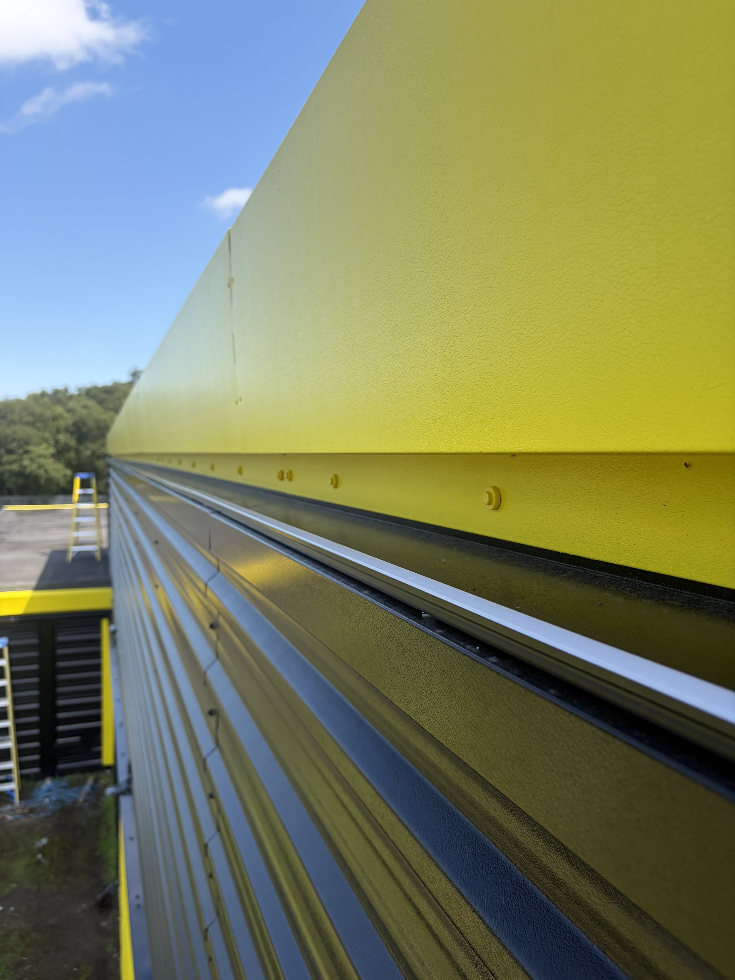 Close-up of a yellow guardrail along a roadway, with cloudy sky and trees in the background.