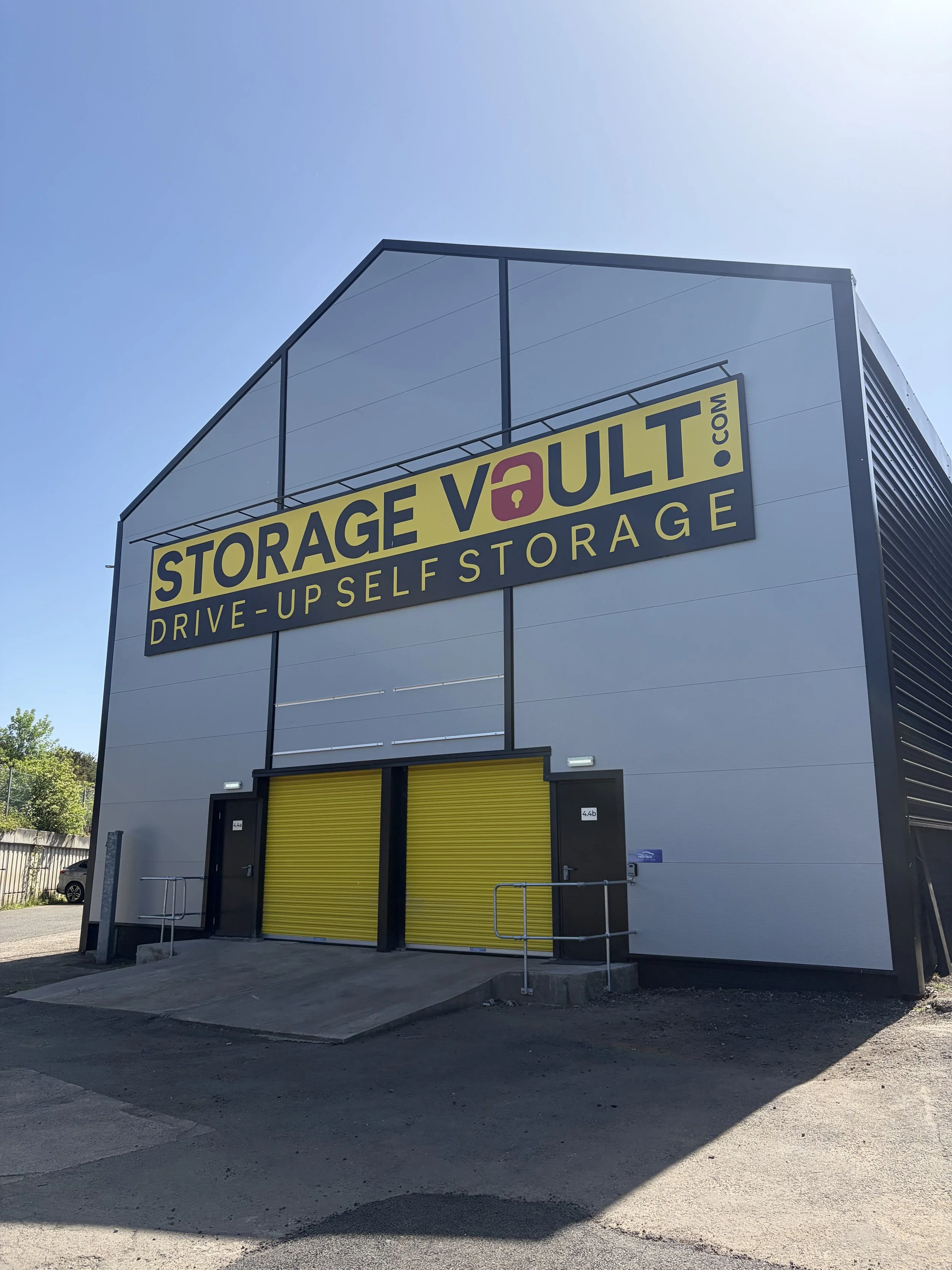 A modern commercial storage facility with yellow roll-up doors and a large yellow sign that reads 'Storage Vault' with a padlock icon and the website 'storagevault.com'. The building has a gray and black exterior with a small ramp leading into the storage units. The sky is clear and sunny.