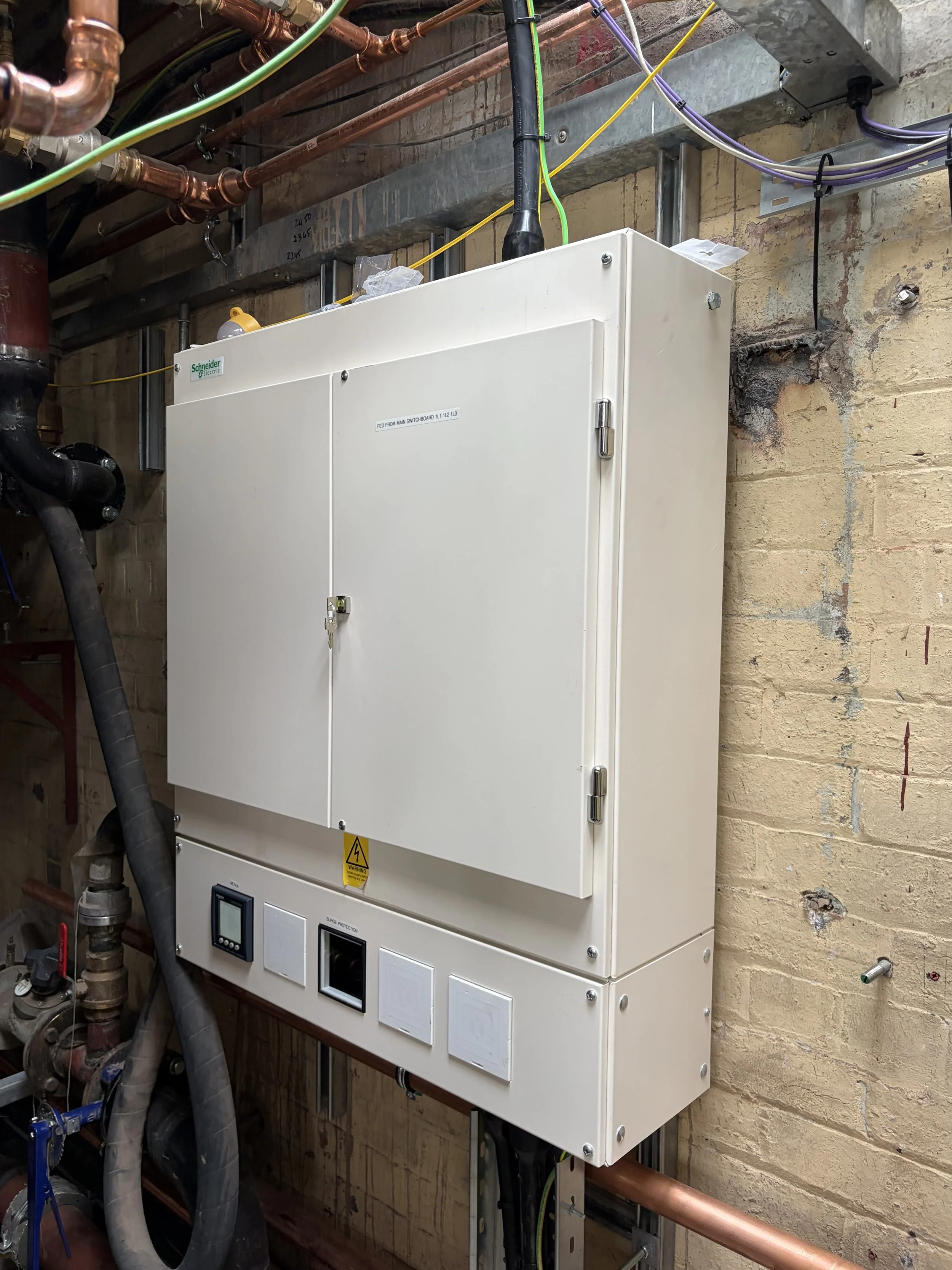 Electrical panel mounted on a brick wall with copper pipes, black pipes, and electrical cables around it.