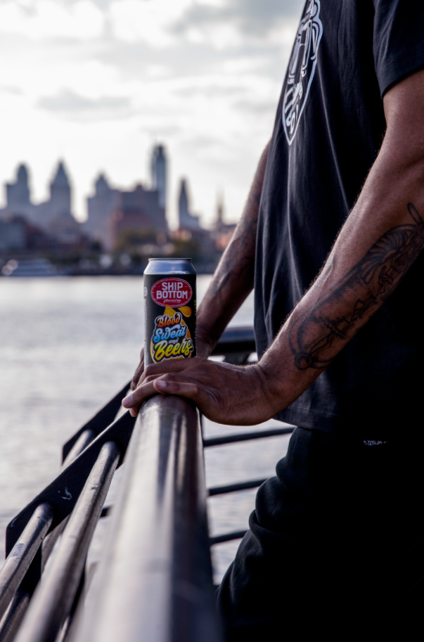 Basketball player Rasheed Wallace with tattoos on his arm, holding a can of 215 Libations/Ship Bottom beer on a railing by the river, with a Philadelphia city skyline in the background.
