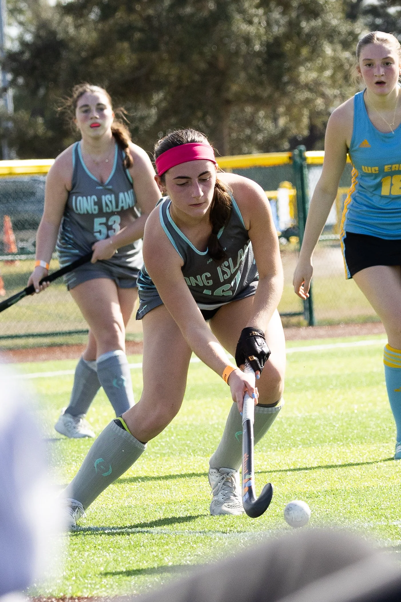 Three young women playing field hockey outdoors. One in the foreground is wearing a gray and teal uniform with a pink headband, about to hit a white ball with her hockey stick. Two others are in the background, also in teal and gray uniforms, on a gr