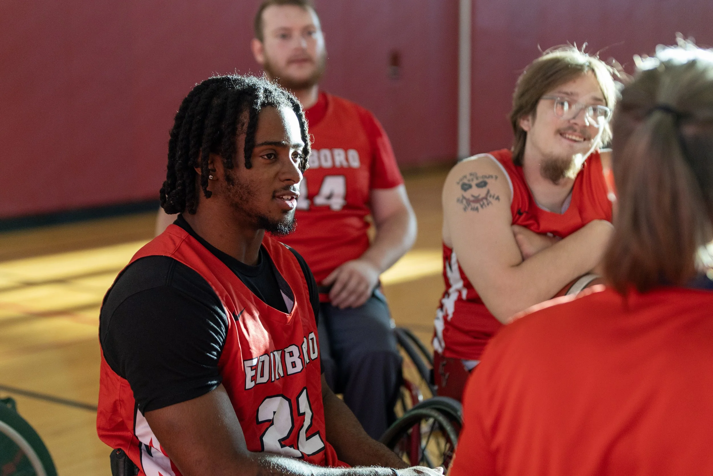 Group of young adults, some in wheelchairs, wearing red sports jerseys, engaged in conversation in a gymnasium. Basketball. Athlete and Sports Photography