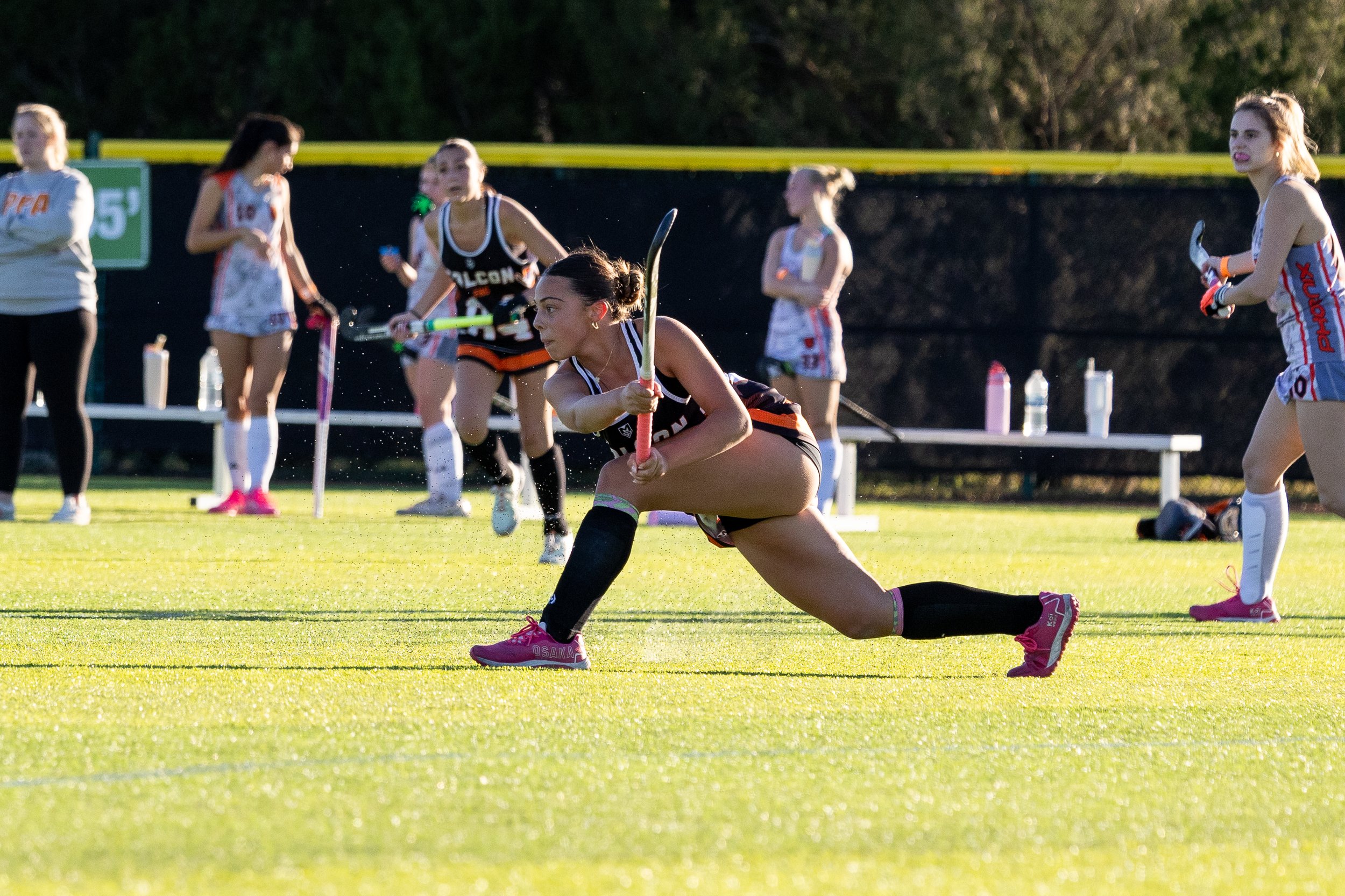 Female field hockey player in black uniform crouching with stick on grass field, surrounded by other players and a coach, during a game.Athlete and Sports Photography