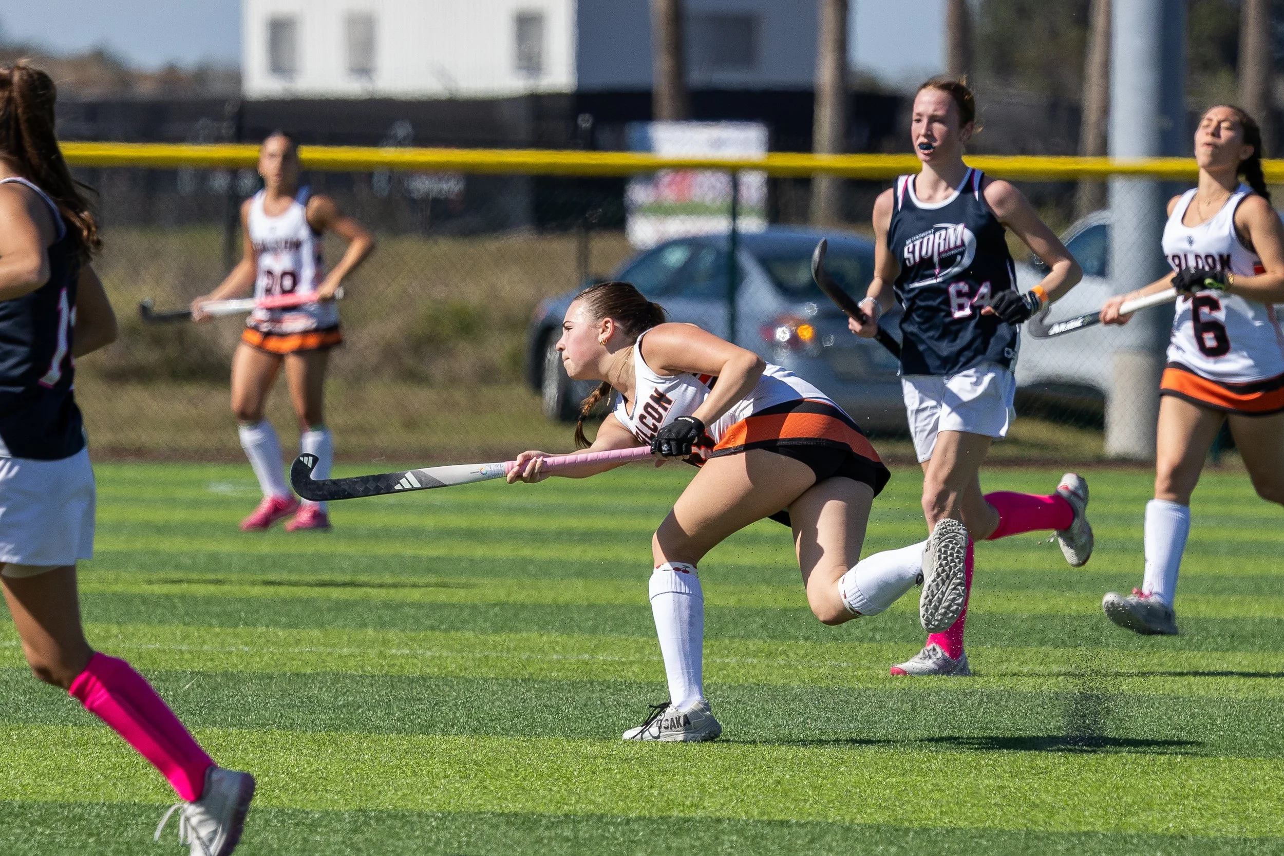 Women's field hockey game with players in action, running on a grassy field, holding hockey sticks, wearing athletic uniforms, and athletic shoes, with a background of cars and buildings.