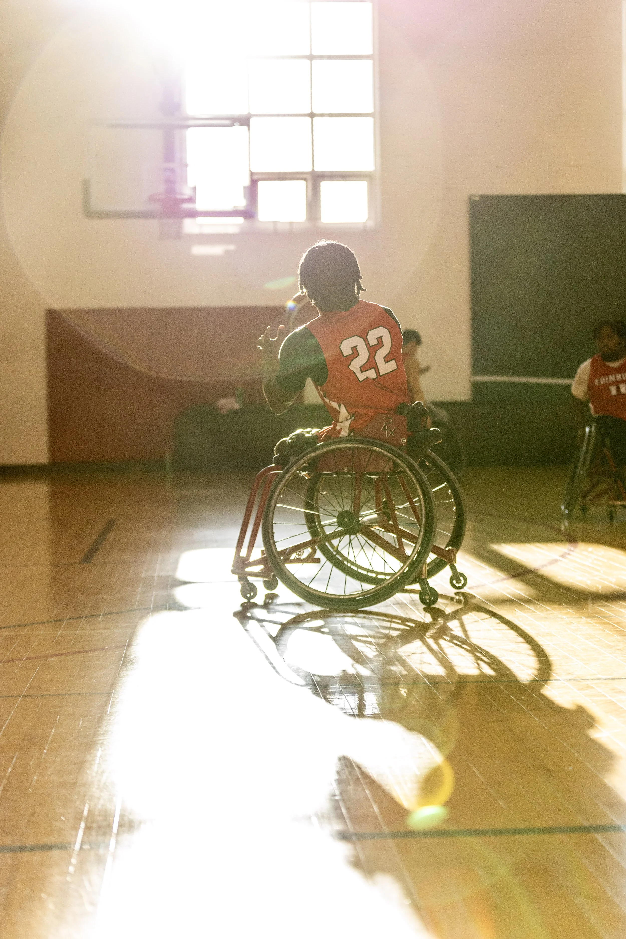 A person in a wheelchair playing basketball indoors, sunlight streaming through a window and casting a shadow on the floor. Athlete and Sports Photography