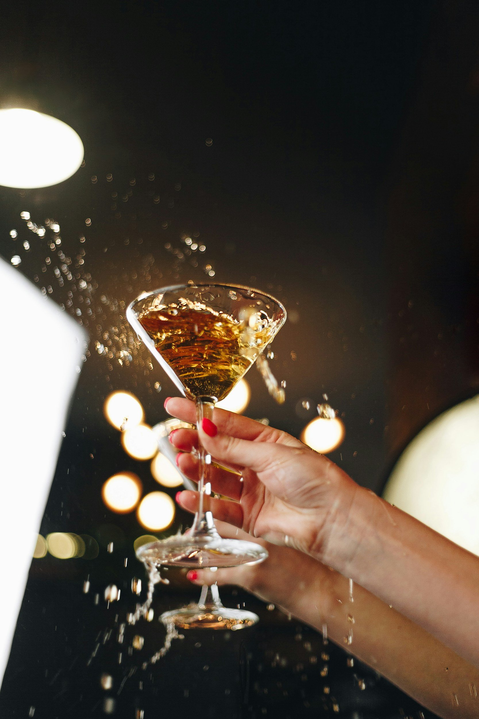 Person holding a cocktail glass with a drink, splashing the liquid, against a dark, blurred background with circular lights.