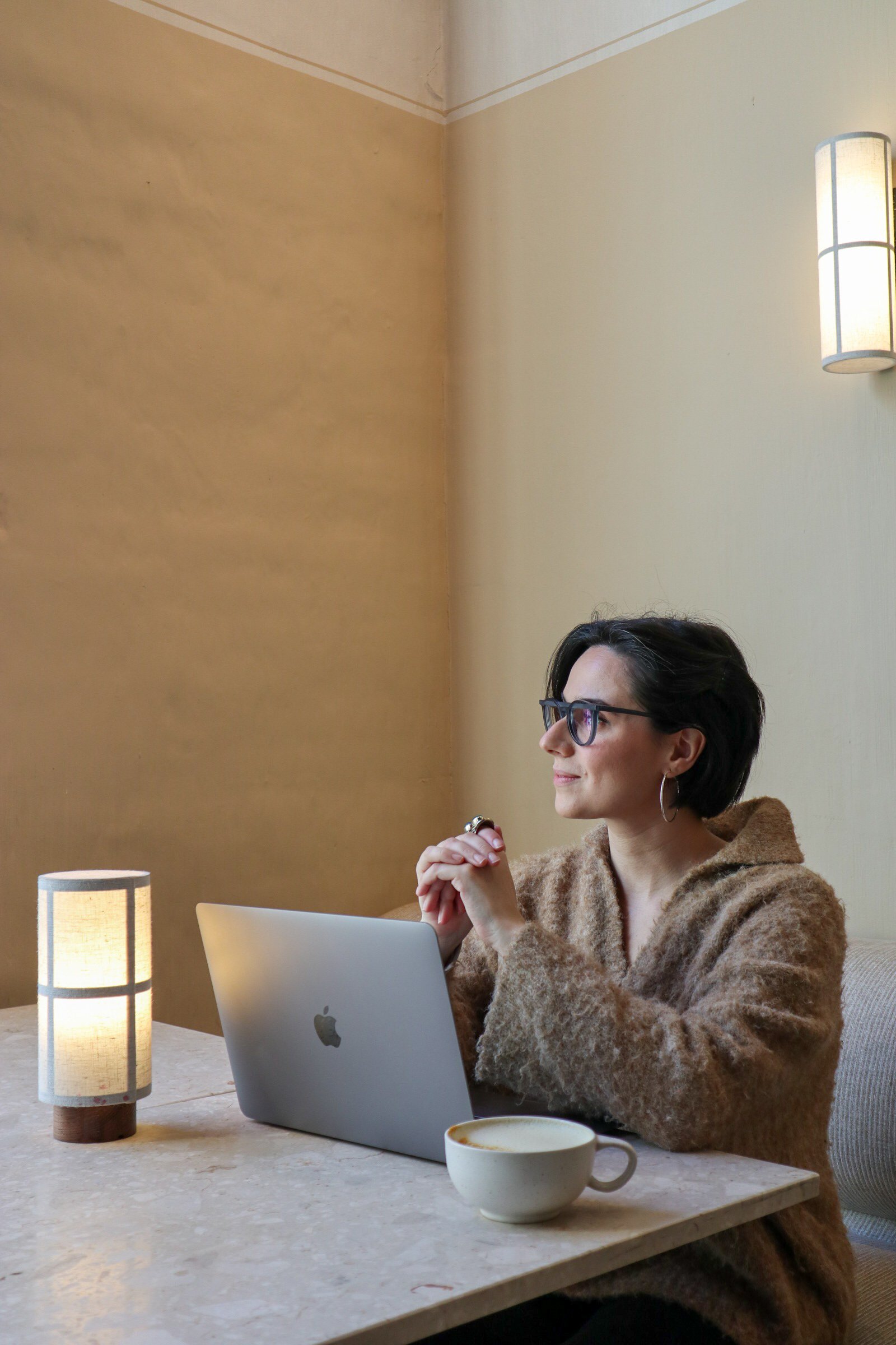 A woman with short dark hair, glasses, and hoop earrings sits at a marble table with a MacBook, a coffee cup, and a lamp, smiling thoughtfully in a warmly lit room.