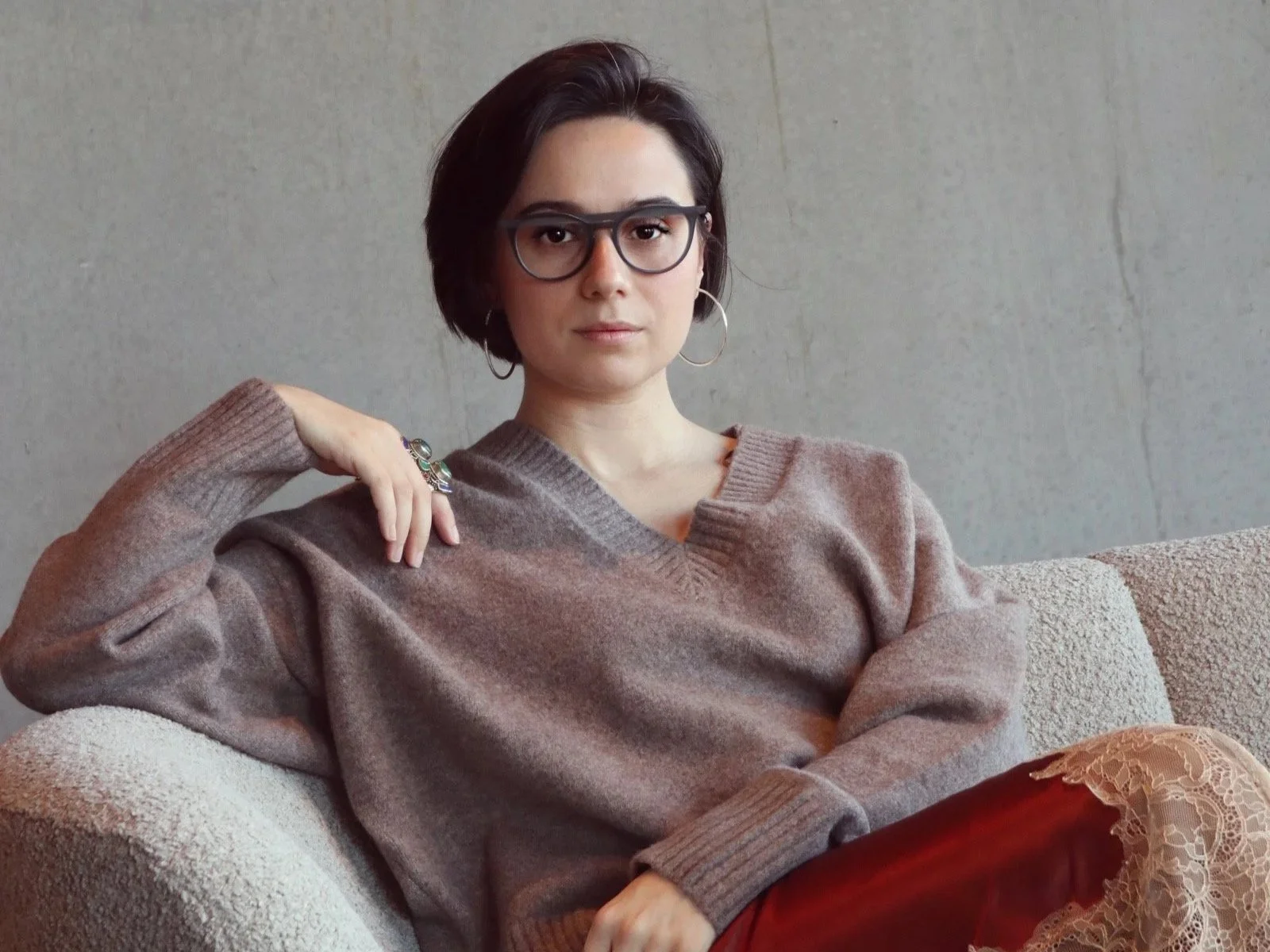 A young woman with short dark hair, glasses, and earrings sitting on a couch against a plain background, wearing a brown sweater and a lace skirt.