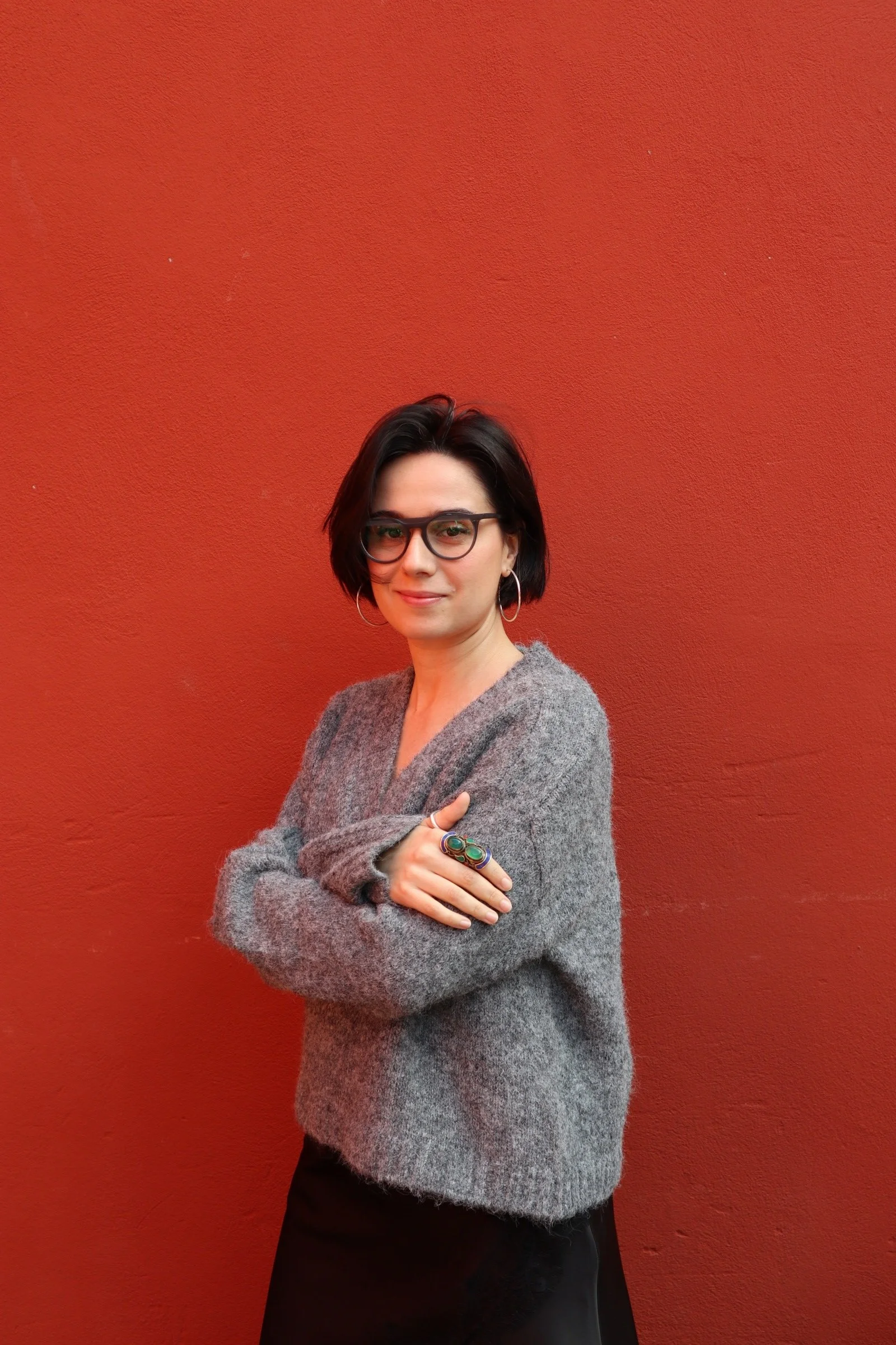 A woman with short black hair, wearing glasses, hoop earrings, a gray sweater, and a dark skirt, standing with arms crossed in front of a reddish-orange wall.