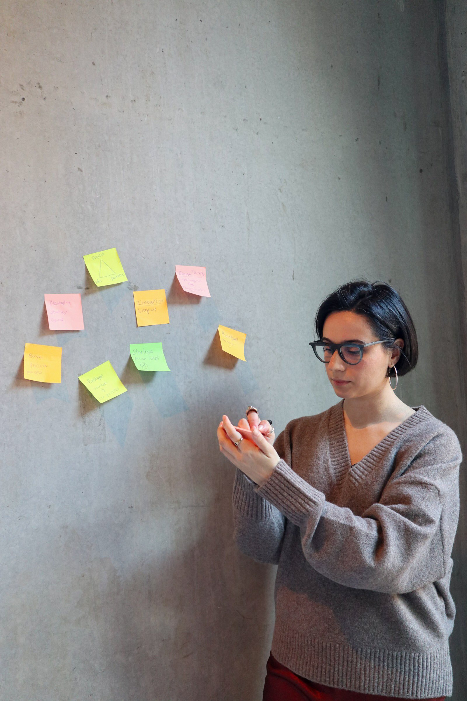 Woman with short dark hair, glasses, wearing a brown sweater, standing in front of a gray wall with colorful sticky notes arranged in a pattern.