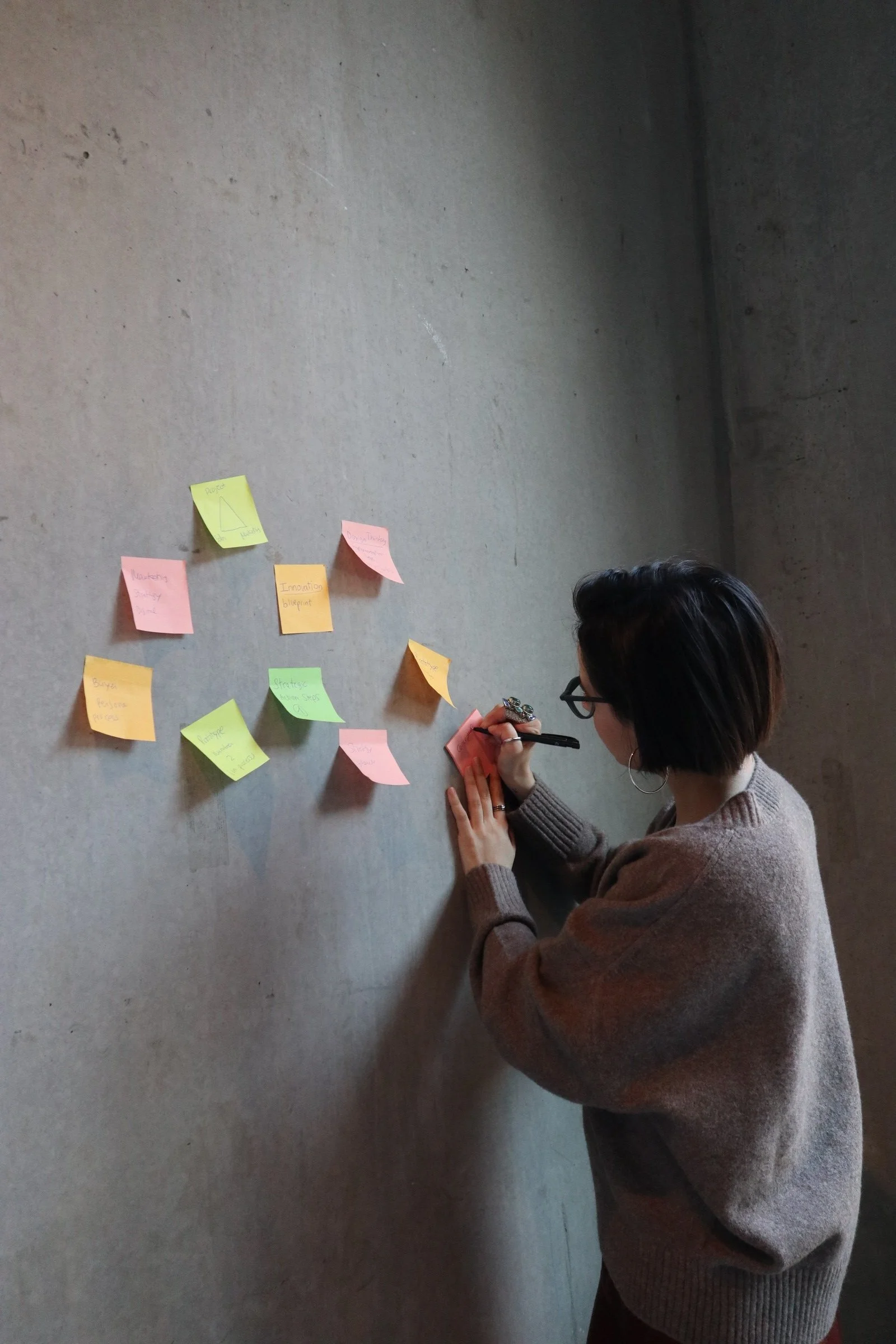 A woman with short dark hair and glasses writing on a pink sticky note on a concrete wall with other colorful sticky notes nearby.