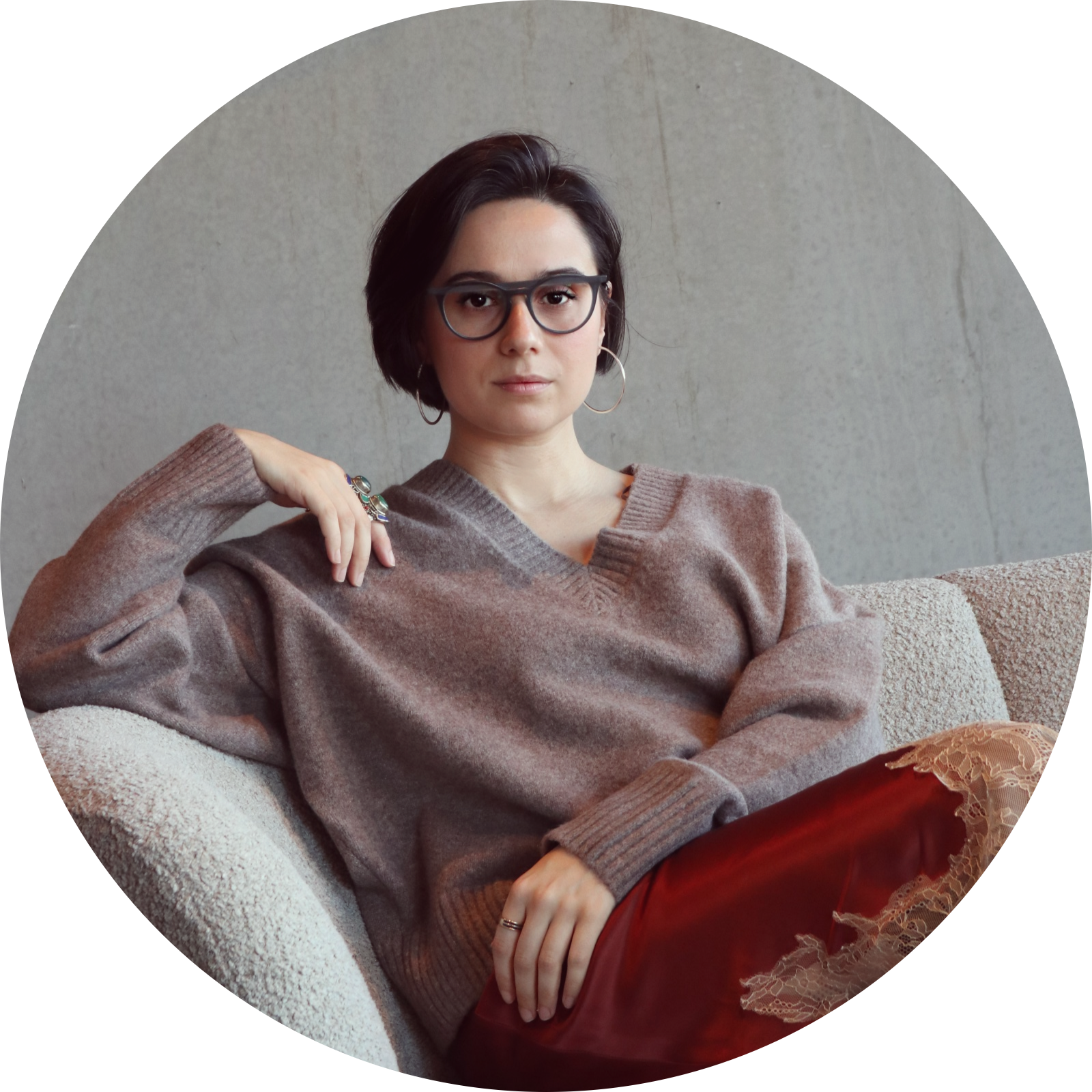 A young woman with short dark hair, wearing glasses, hoop earrings, a brown V-neck sweater, and a shiny red skirt with lace trim, sitting on a beige textured sofa in front of a plain gray wall.
