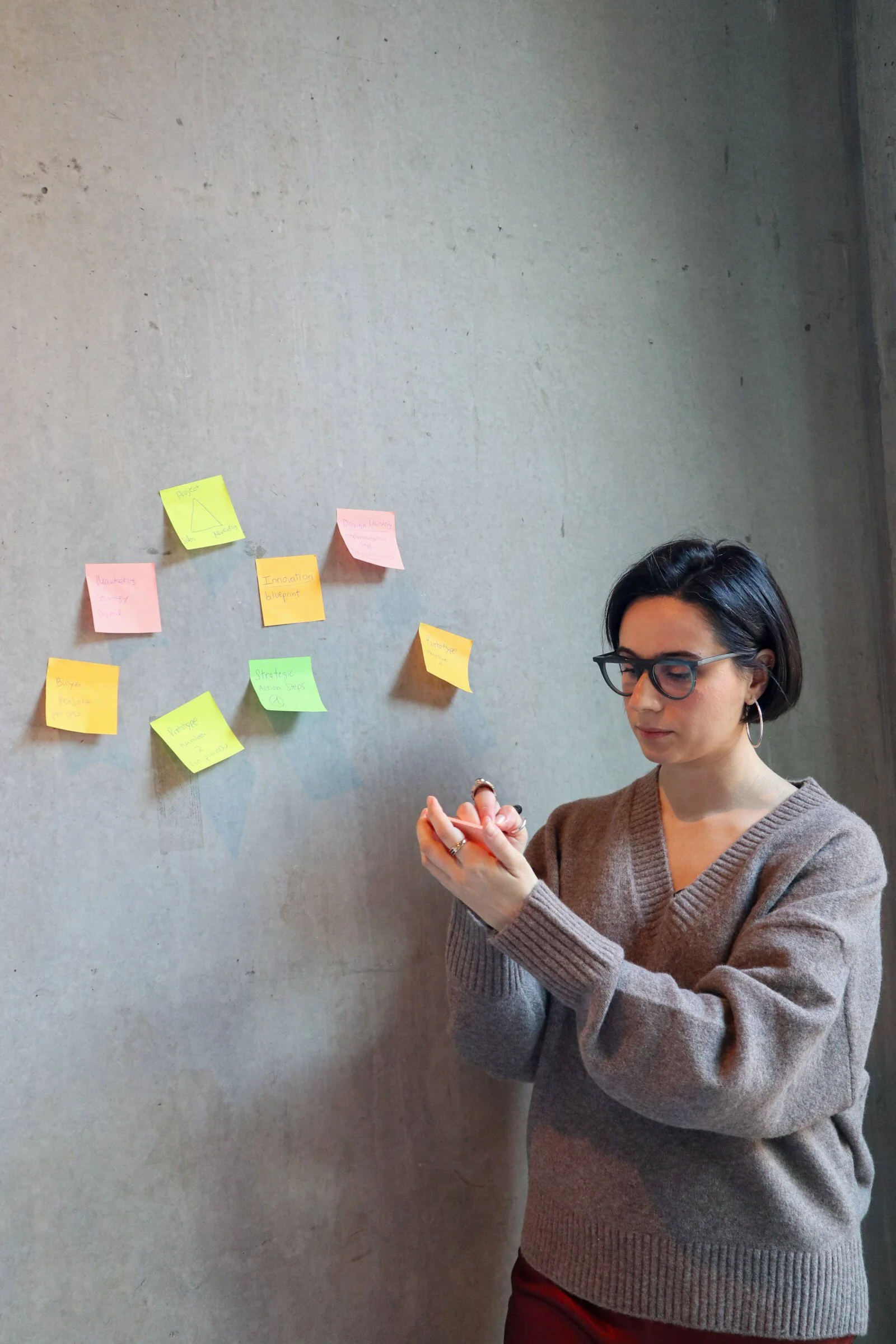 A woman with short black hair, glasses, hoop earrings, wearing a brown sweater, is writing on a notepad while standing in front of a concrete wall with colorful sticky notes.