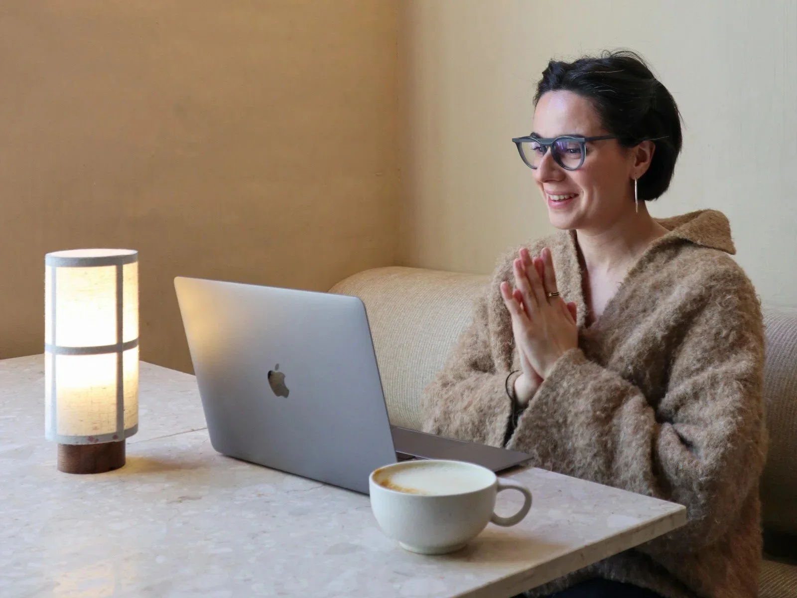 A woman with short dark hair, glasses, and earrings sits at a table with a MacBook, smiling with hands pressed together in front of her. There is a cup of coffee and a lamp on the table.