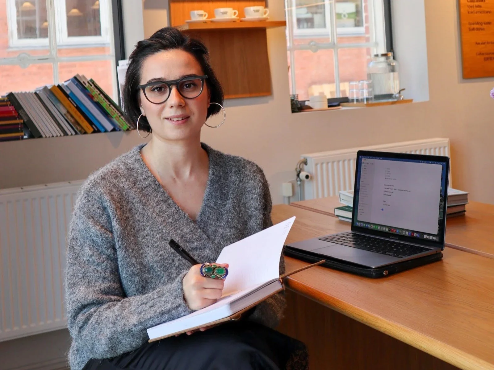 A woman with short dark hair, glasses, and hoop earrings sitting at a wooden table in a cozy cafe, holding a pen and notebook, with a laptop open beside her.