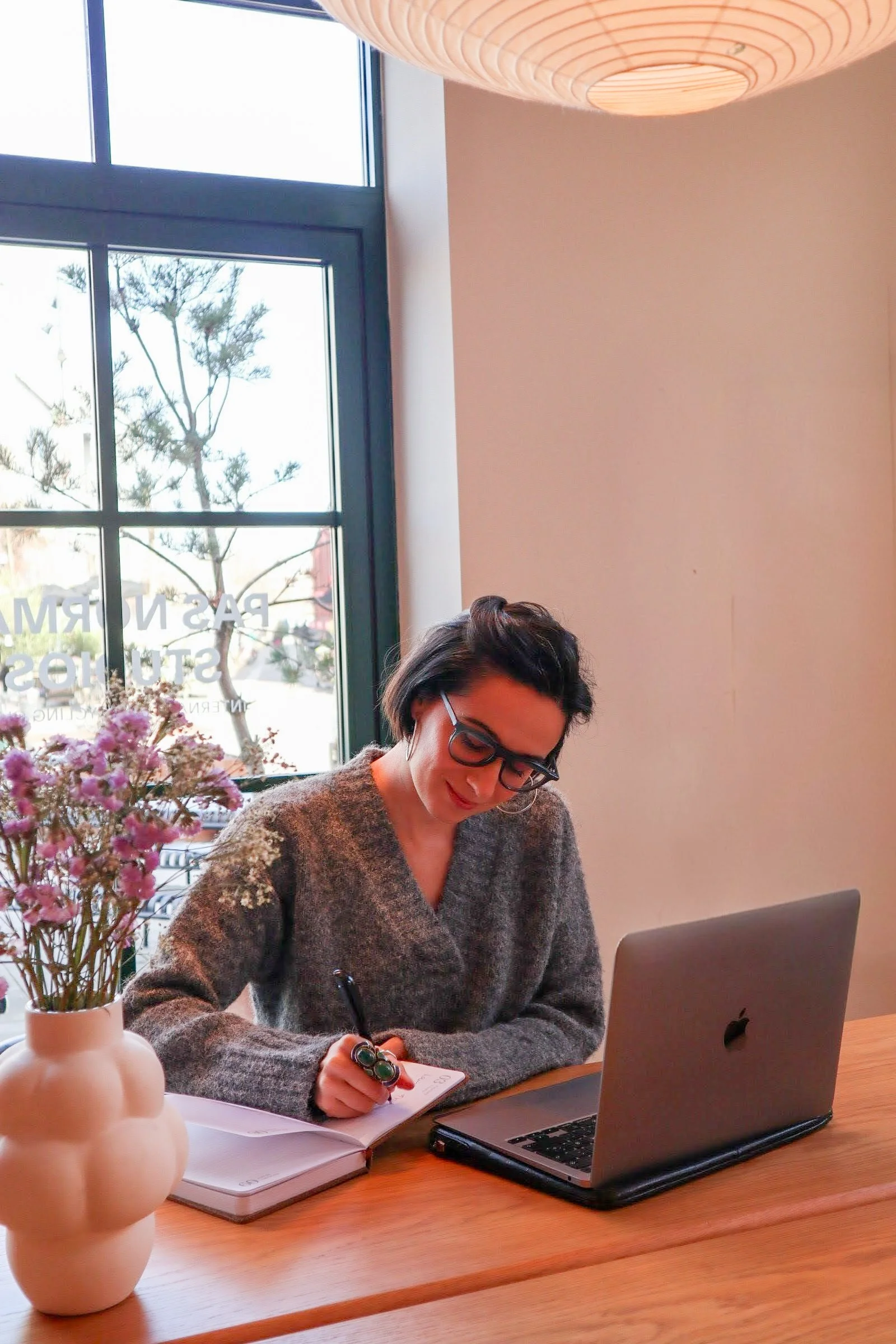 A woman with short dark hair and glasses, sitting at a wooden table, writing in a notebook with a pen, with a laptop and a vase of pink flowers, near a large window in a well-lit room.