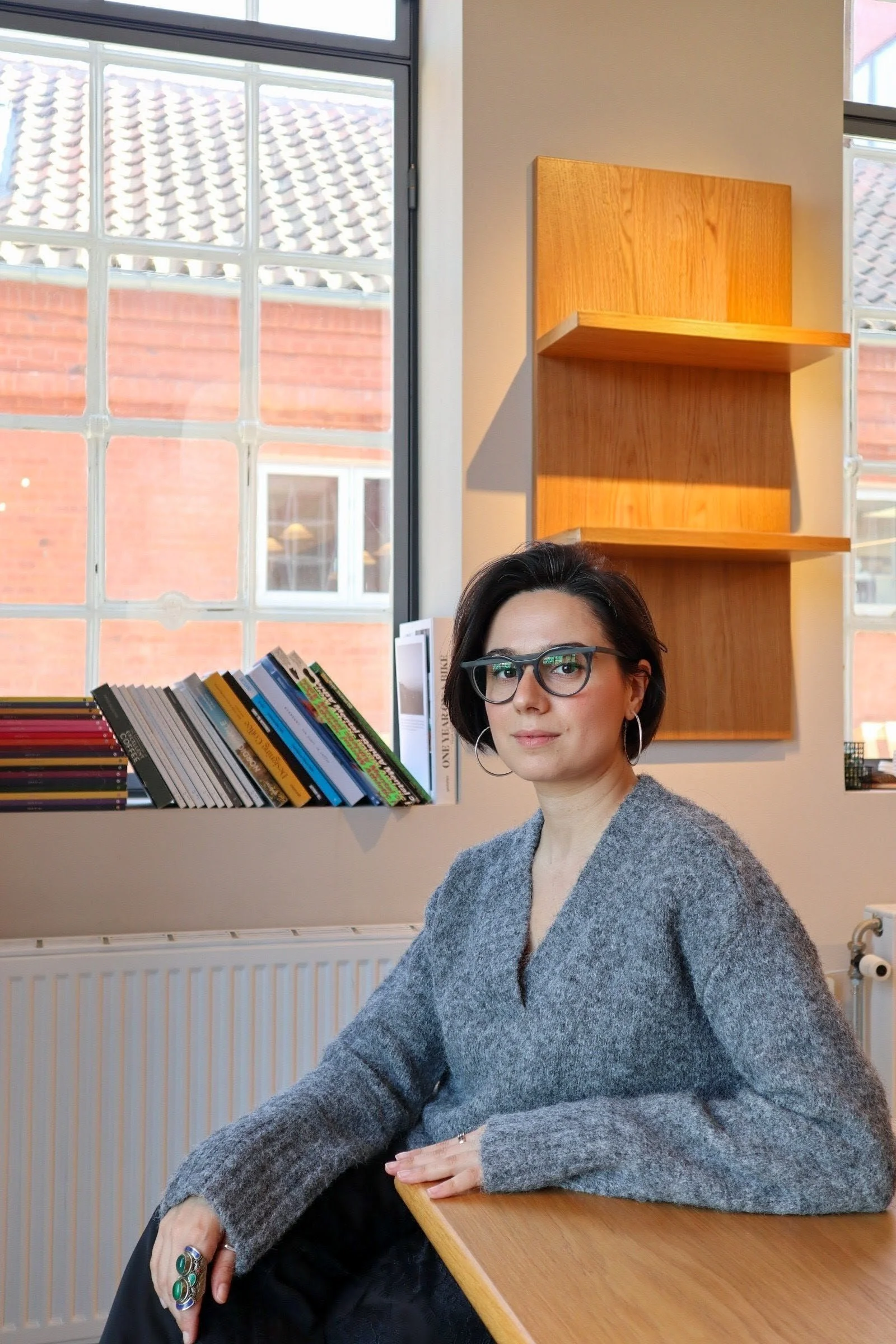 A woman with dark hair, glasses, and hoop earrings sitting at a wooden table in a room with large windows, books on the windowsill, and wooden wall shelves behind her.