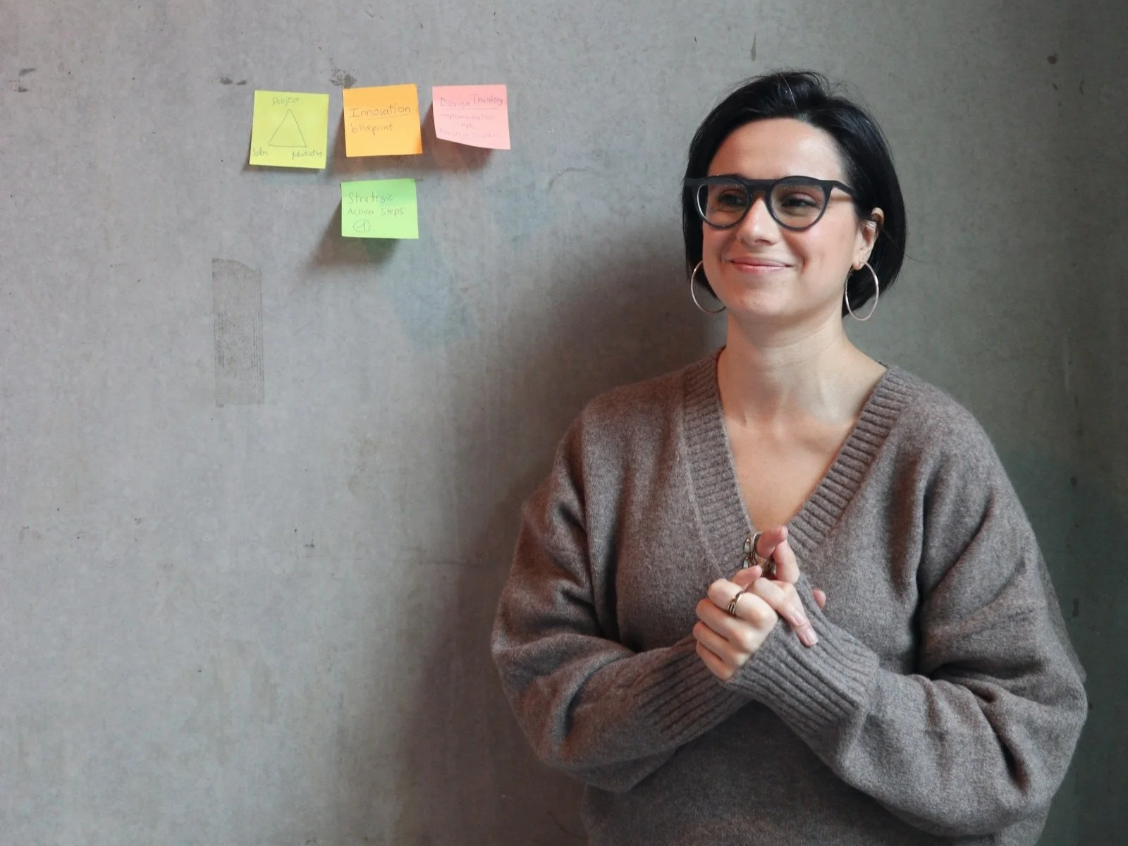 A woman with black hair, glasses, and hoop earrings standing in front of a concrete wall with sticky notes arranged above her head. She is wearing a brown sweater and has a pleasant, thoughtful expression.