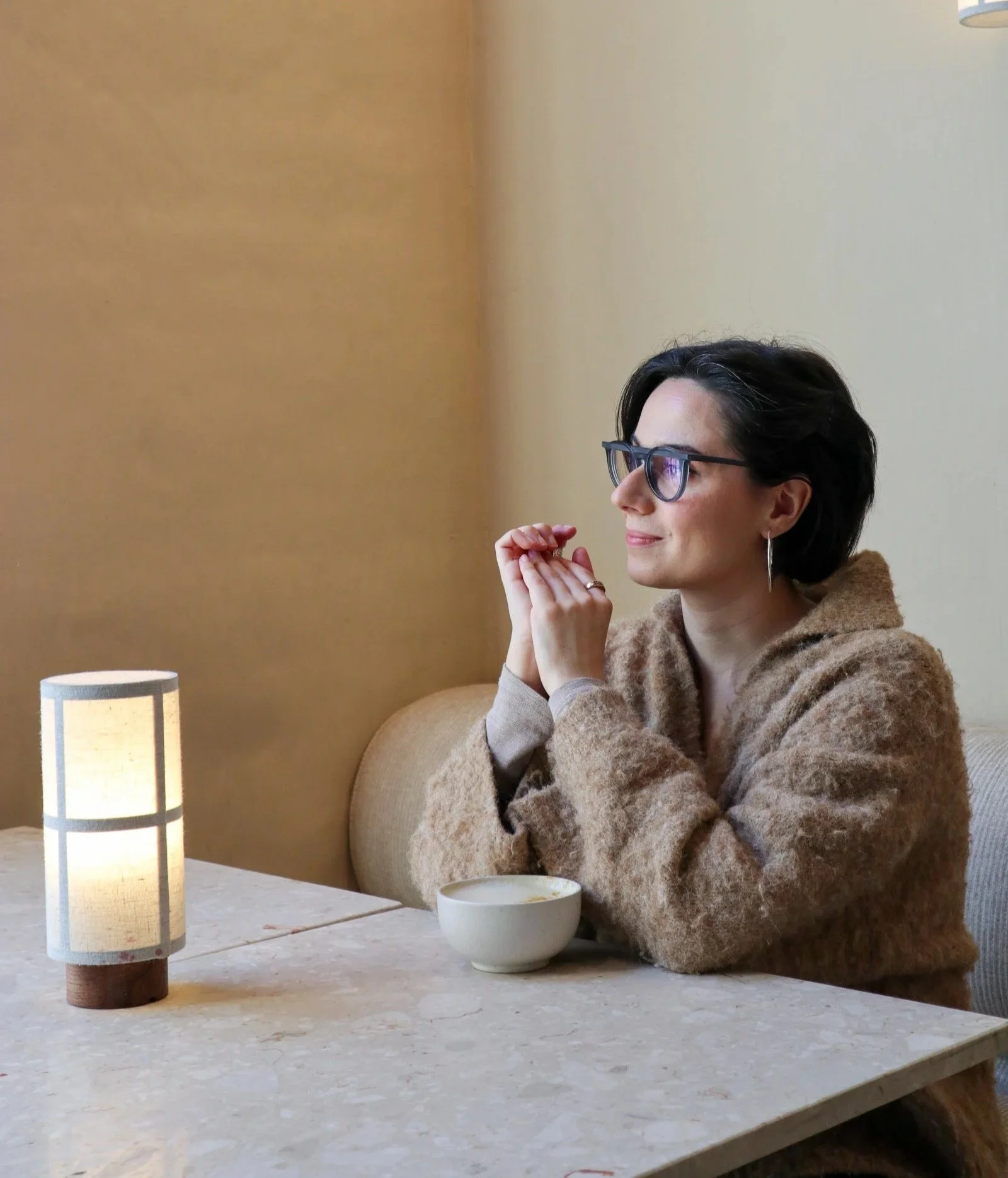 A woman with short dark hair, glasses, and hoop earrings sitting at a table with her hands clasped, in a cozy restaurant or cafe. There is a cup of coffee or tea in front of her, and a small lamp on the table providing warm light. She is wearing a brown fuzzy sweater.