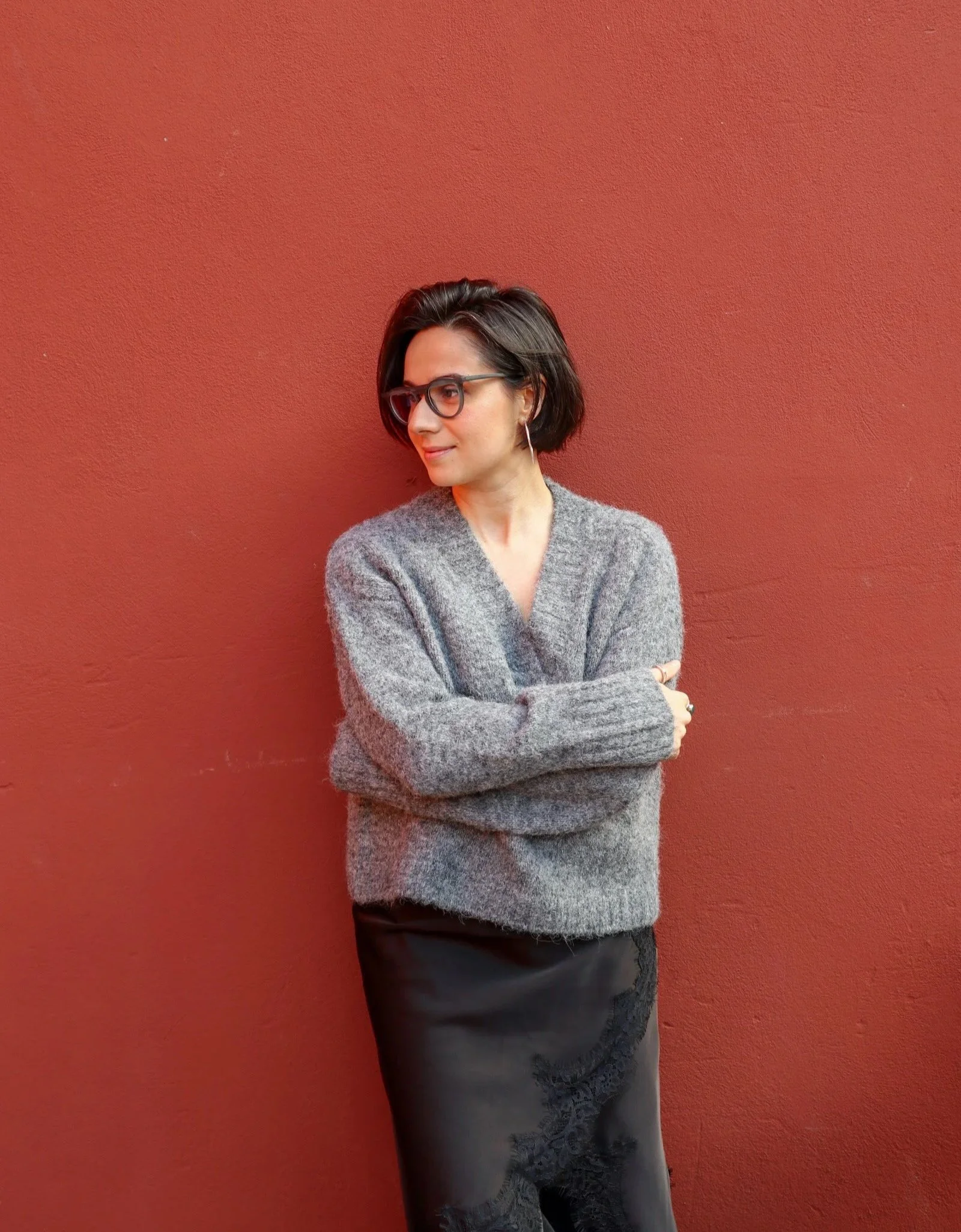 Woman with short dark hair and glasses, wearing a gray sweater and black skirt, standing against a red wall with arms crossed.