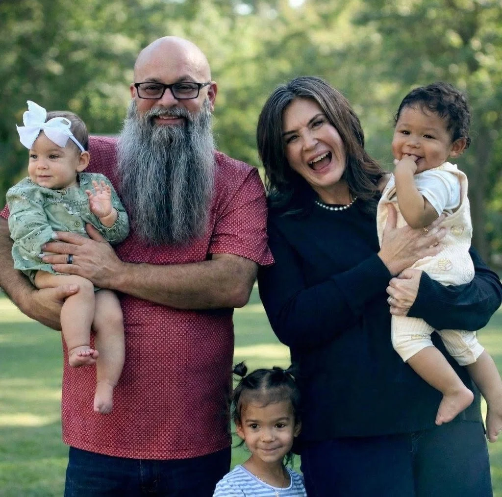 A smiling family poses outdoors, with a man with a beard and glasses, a woman, and three young children, all holding each other on a grassy area with trees in the background.