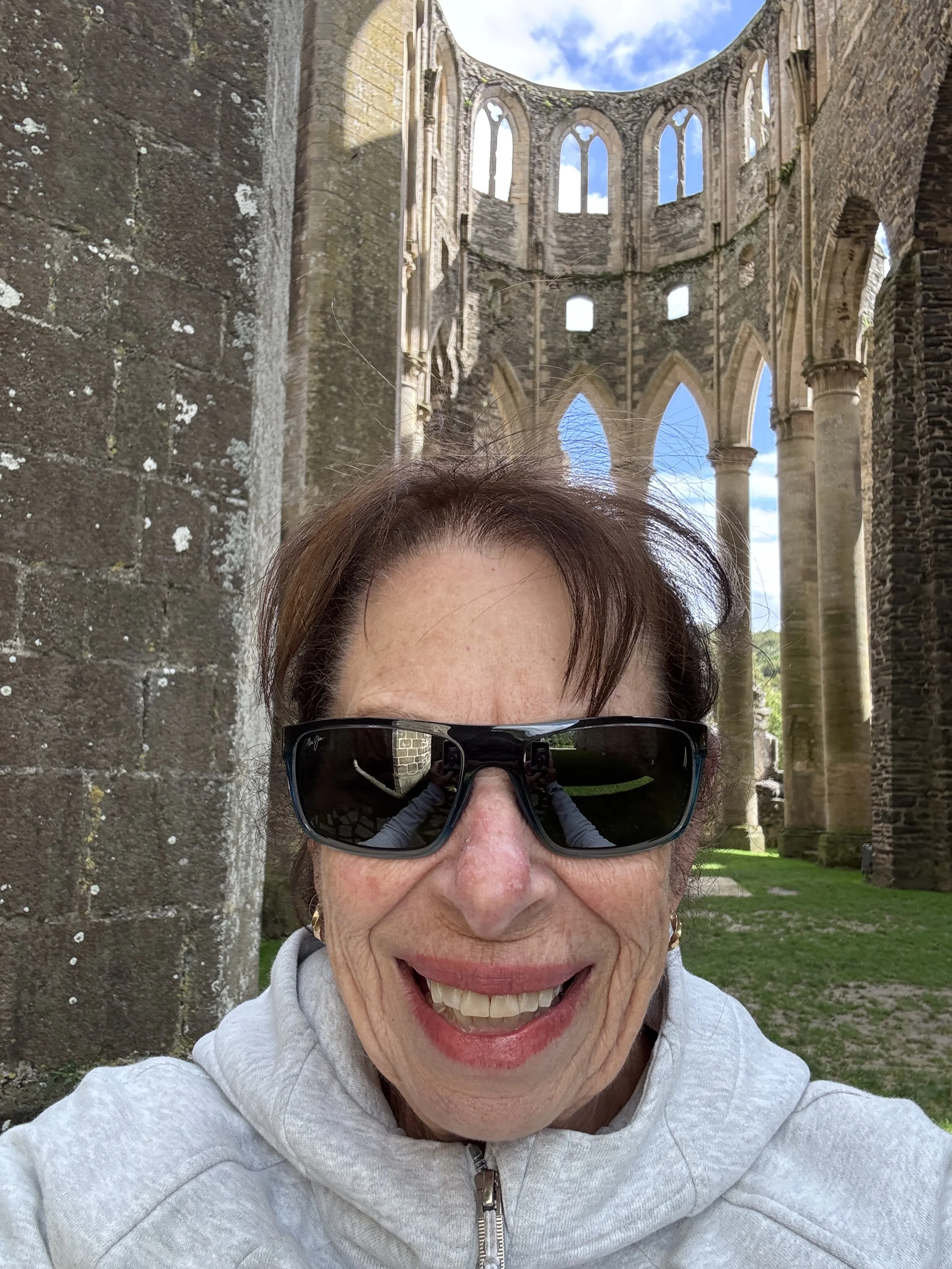 A smiling woman wearing sunglasses and a light-colored hoodie, taking a selfie in front of the ruins of an old stone church with tall, arched windows and columns.
