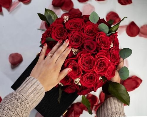 A person holding a bouquet of red roses with green leaves, surrounded by scattered rose petals on a white surface.
