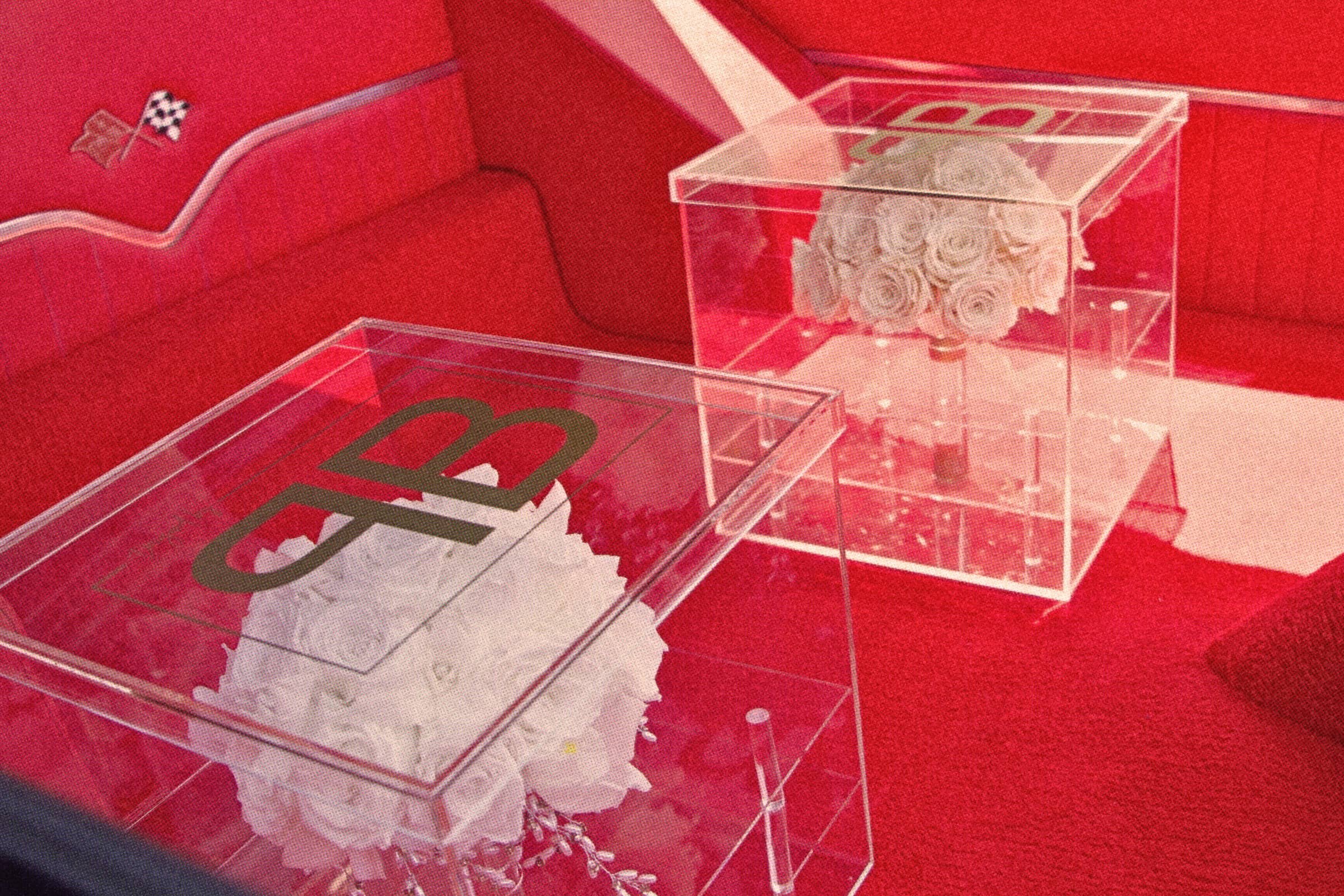 Two clear acrylic boxes containing white flower bouquets, placed on a red surface, with a red background and a brown couch behind them.