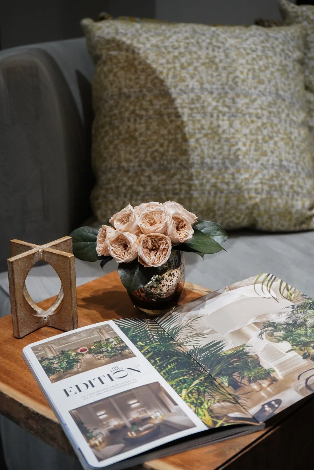 A wooden side table with an open magazine, a small floral arrangement of pale pink roses in a decorative vase, and a gold colored decorative frame. In the background, a sofa with a large yellow and white patterned cushion.