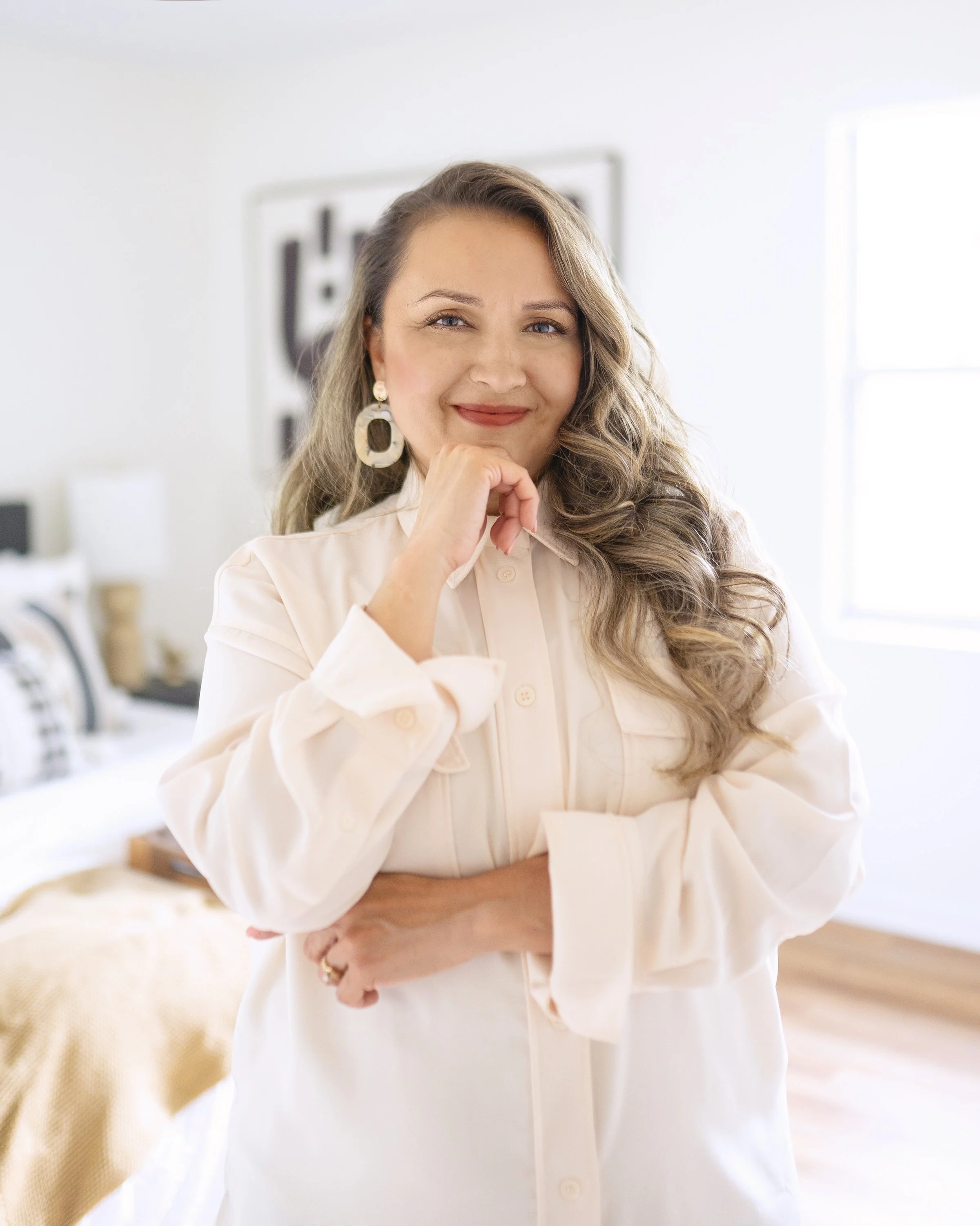 A woman with long, wavy hair smiling and resting her chin on her hand, standing in a bright, modern bedroom with a framed artwork and window in the background.