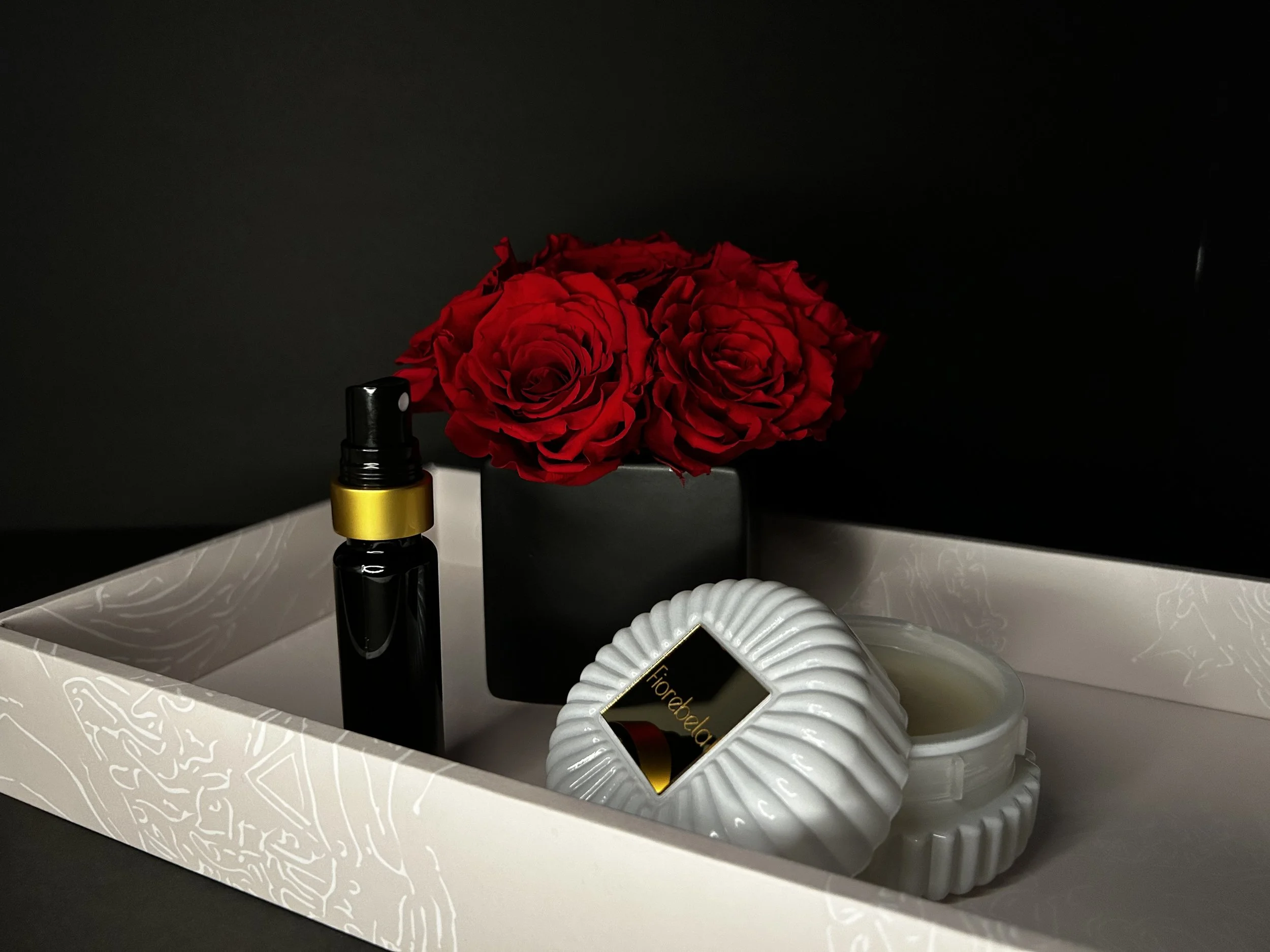 A decorative tray with a small jar of candles, a spray bottle, and a black box with red roses in a black vase.