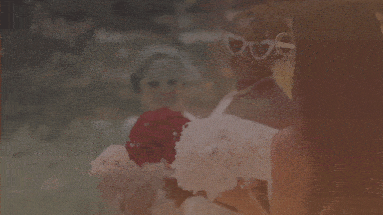 A woman wearing sunglasses and a wide-brimmed hat, holding a bouquet of red and white flowers, with a younger person in the background.