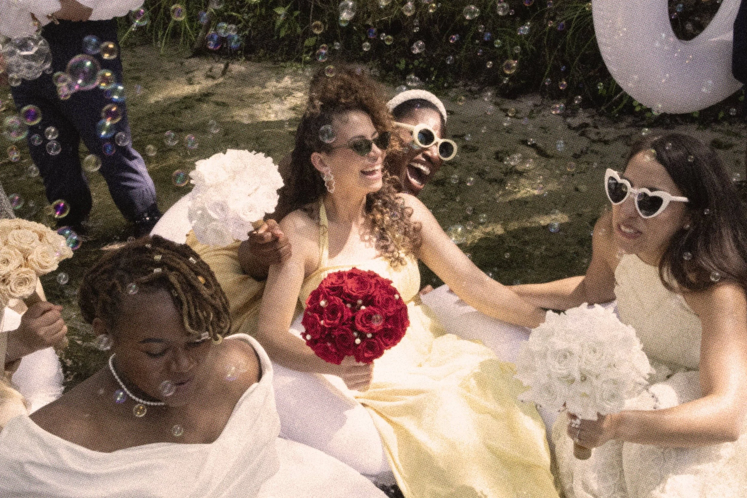 A group of women at a wedding celebration, sitting in a boat, smiling, holding bouquets, some wearing sunglasses, surrounded by bubbles.