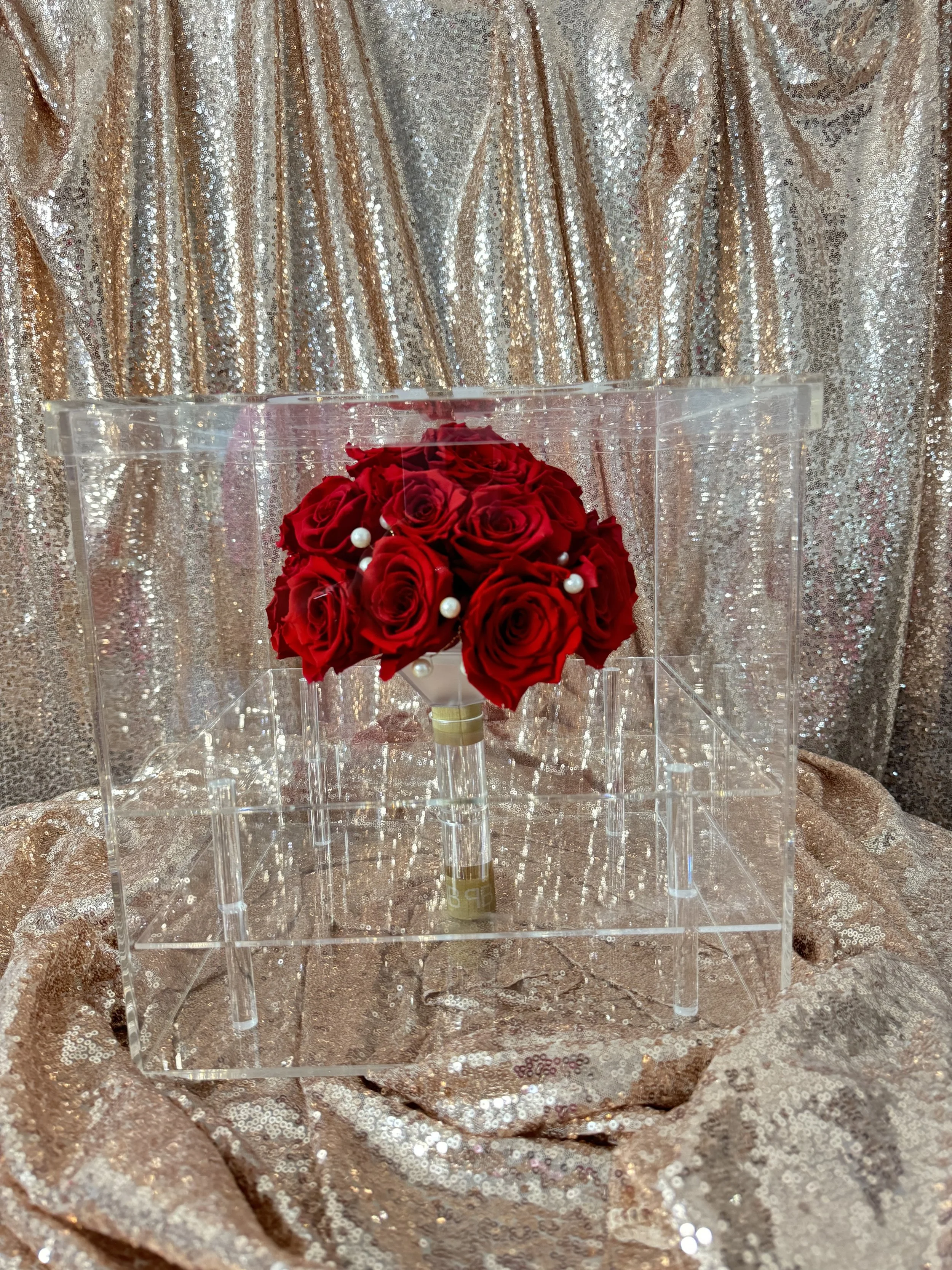 A boutonniere made of red roses with pearl accents, displayed in a clear acrylic case against a glittery gold fabric background.