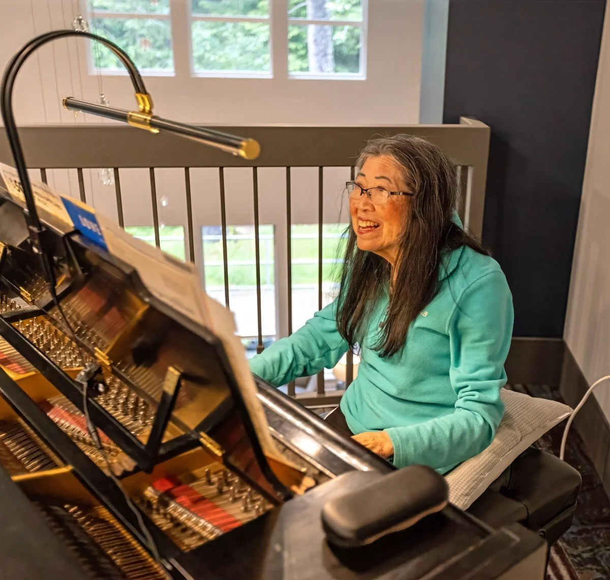 An elderly woman with glasses and long gray hair, wearing a teal shirt, sitting at a grand piano and smiling while playing the piano at Riverview Senior Living and Memory Care in Juneau Alaska