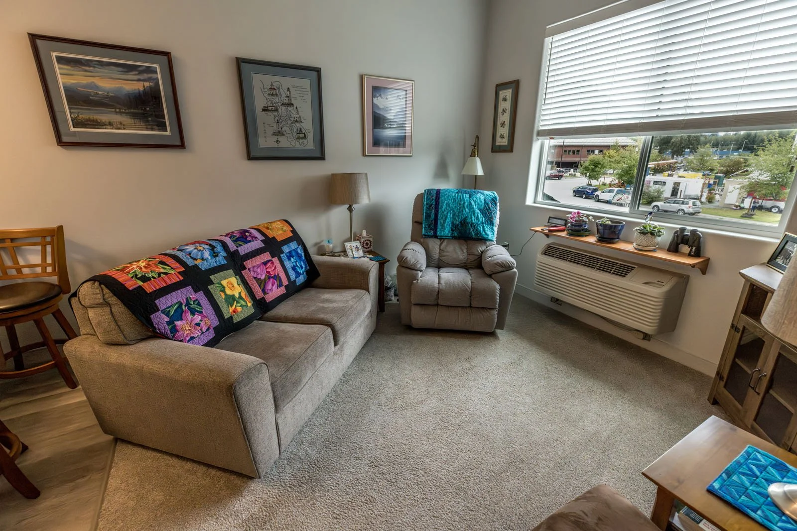 Living room with beige sofa, beige recliner with blue blanket, window with blinds, and wall art at Riverview Senior Living and Memory Care in Juneau Alaska