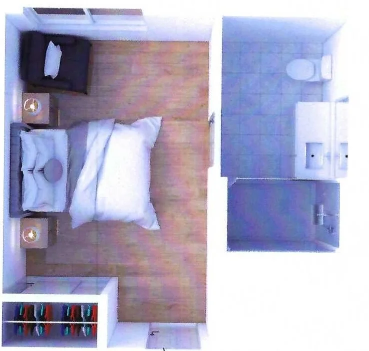Small bedroom with a bed, two side tables, a chair, and an open closet with colorful hangers, adjacent to a kitchenette with a toilet and sink at Riverview Senior Living and Memory Care in Juneau Alaska