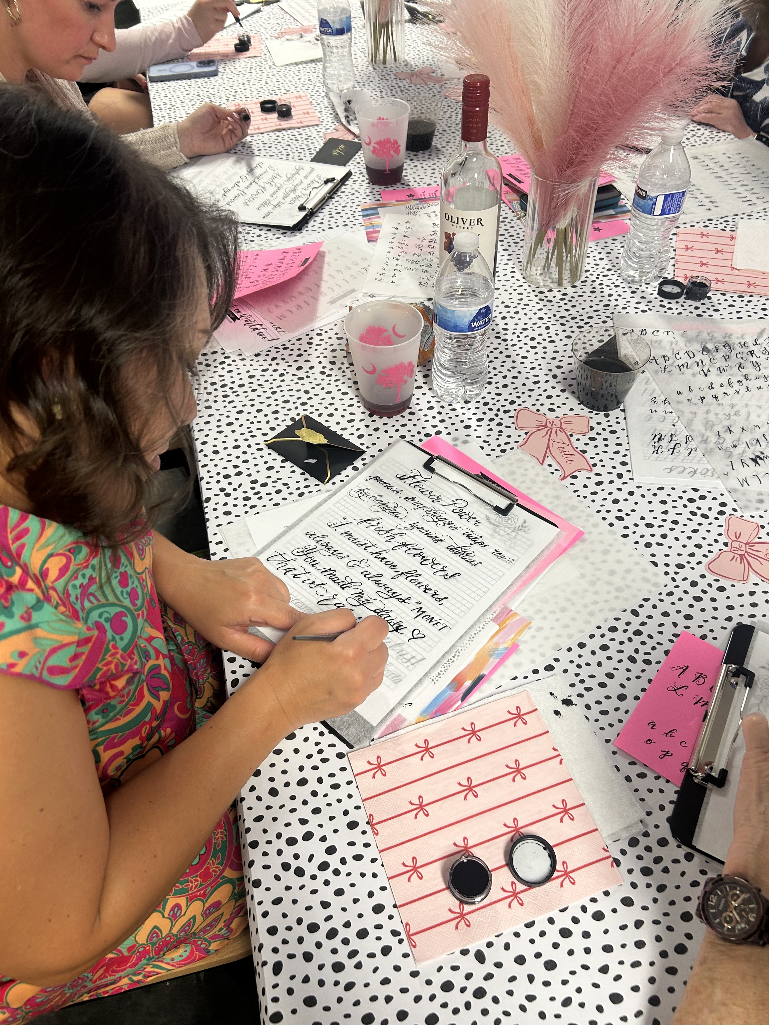 A woman is sitting at a table, writing calligraphy on a sheet of paper. The table is decorated with pink and black themed items, including water bottles, cups, printed papers, and a vase with pink pampas grass. The woman is wearing a colorful, patterned top.