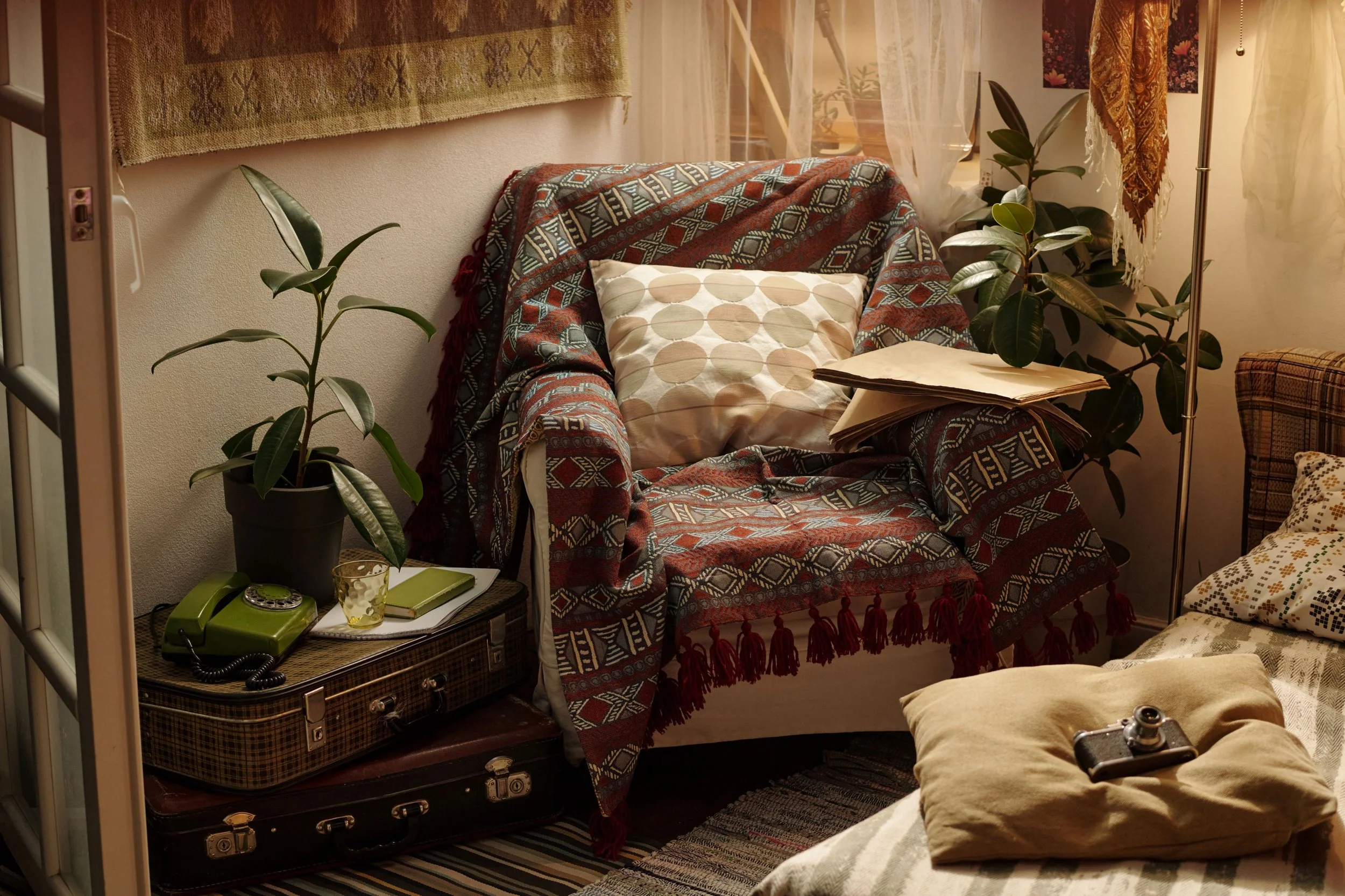 A cozy living room corner with an armchair covered by a patterned blanket, a decorative pillow, and surrounded by potted plants. There are vintage suitcases stacked near the armchair, a green rotary phone, a glass with a drink, and some papers on the side table. A floor lamp and curtains are also visible.