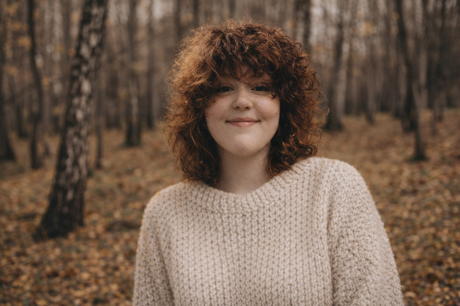 A woman with curly red hair smiling outdoors in a forest with fallen leaves on the ground.