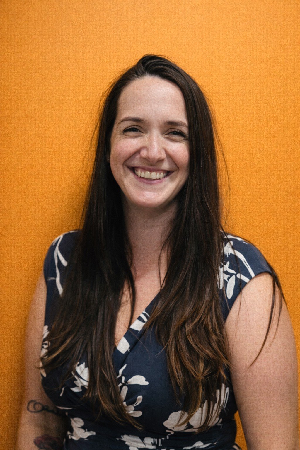 Smiling woman with long dark hair wearing a navy blue and white floral dress standing against an orange wall.