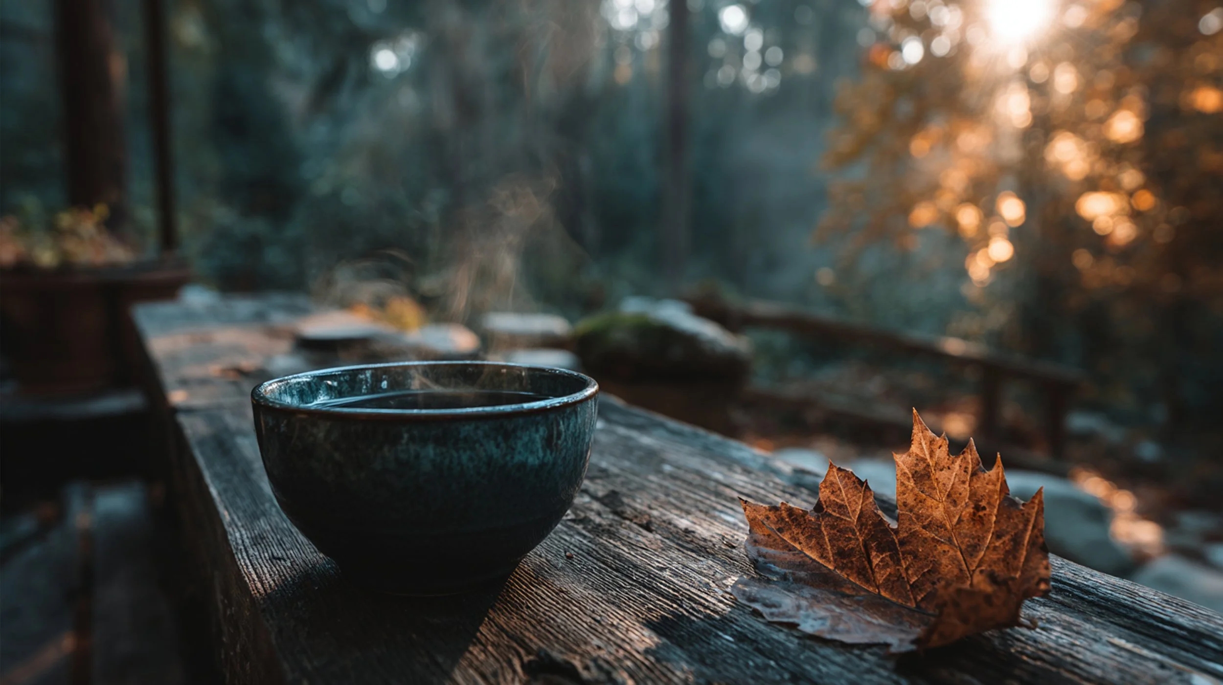 A steaming cup of hot beverage on a rustic wooden table outdoors in a forest setting during sunset, with a dried autumn leaf beside the cup.