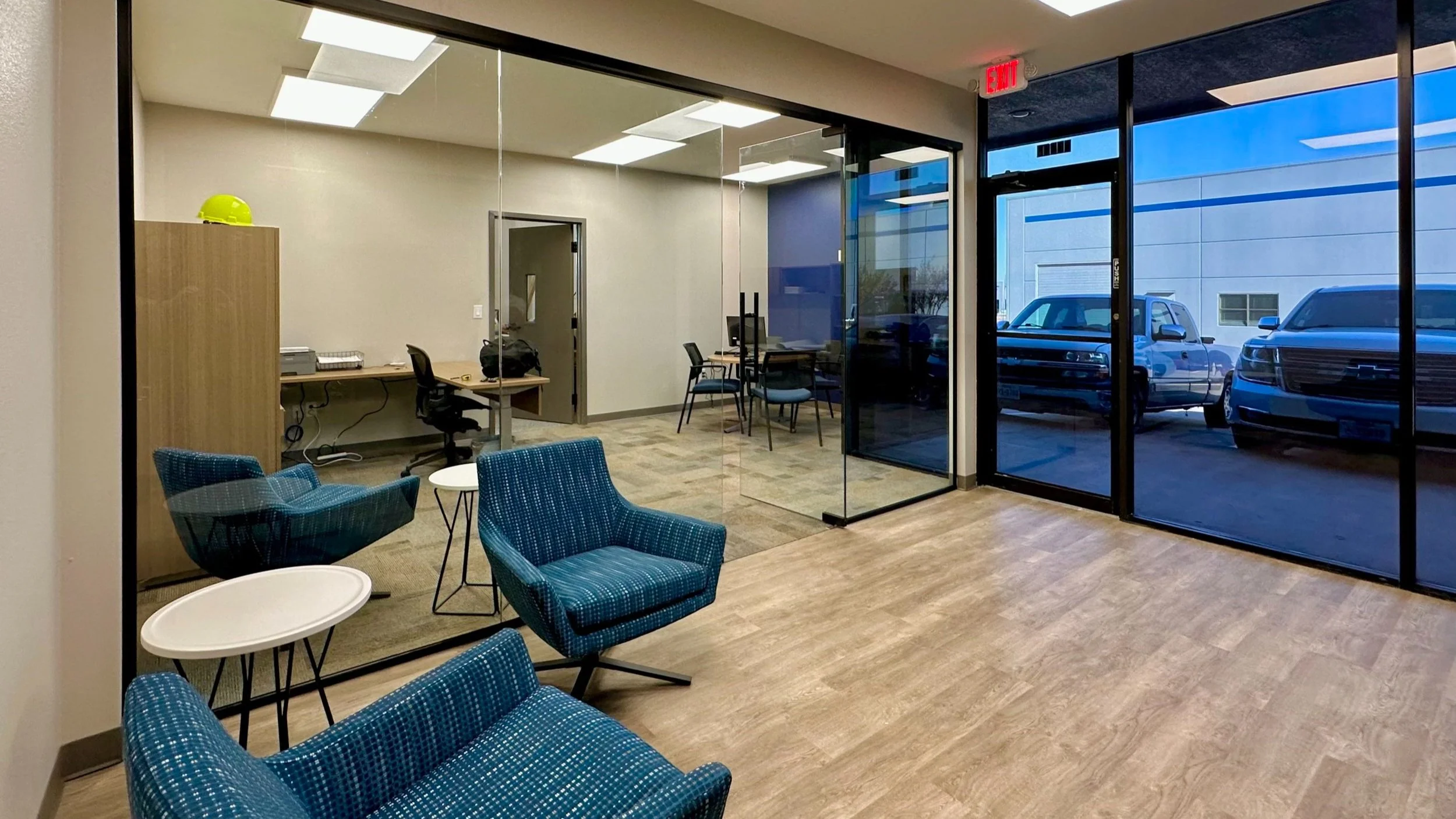 An office lobby with blue patterned chairs and small white tables, a glass-walled meeting room, and a view of parked vehicles outside.